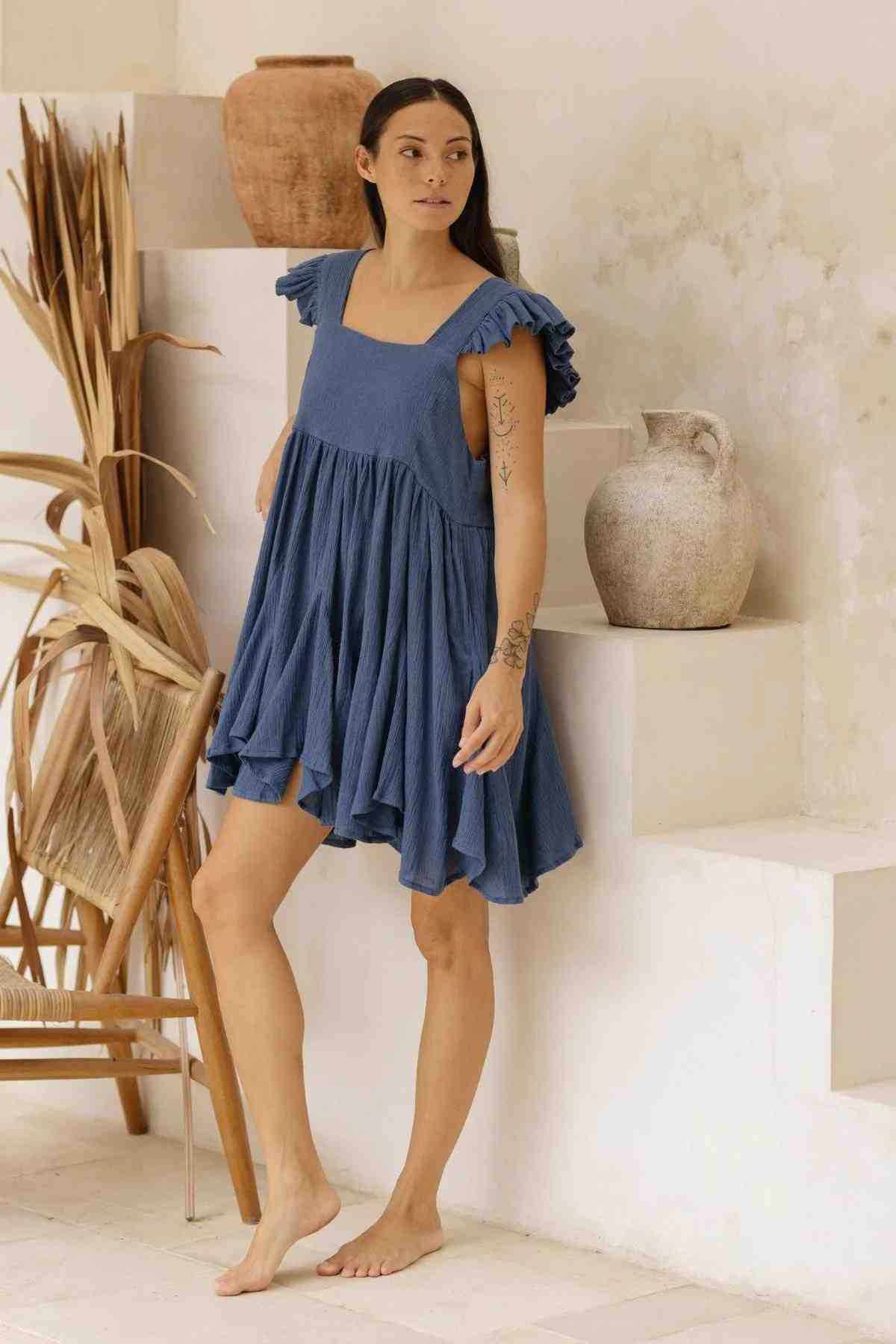 A woman in a blue Anna Yemaya Dress (BGN) by Myrah Penaloza stands barefoot on a white step in a minimalist room. She is surrounded by decorative pottery and dried plants, with the crinkle cotton of her dress adding texture to the serene scene.