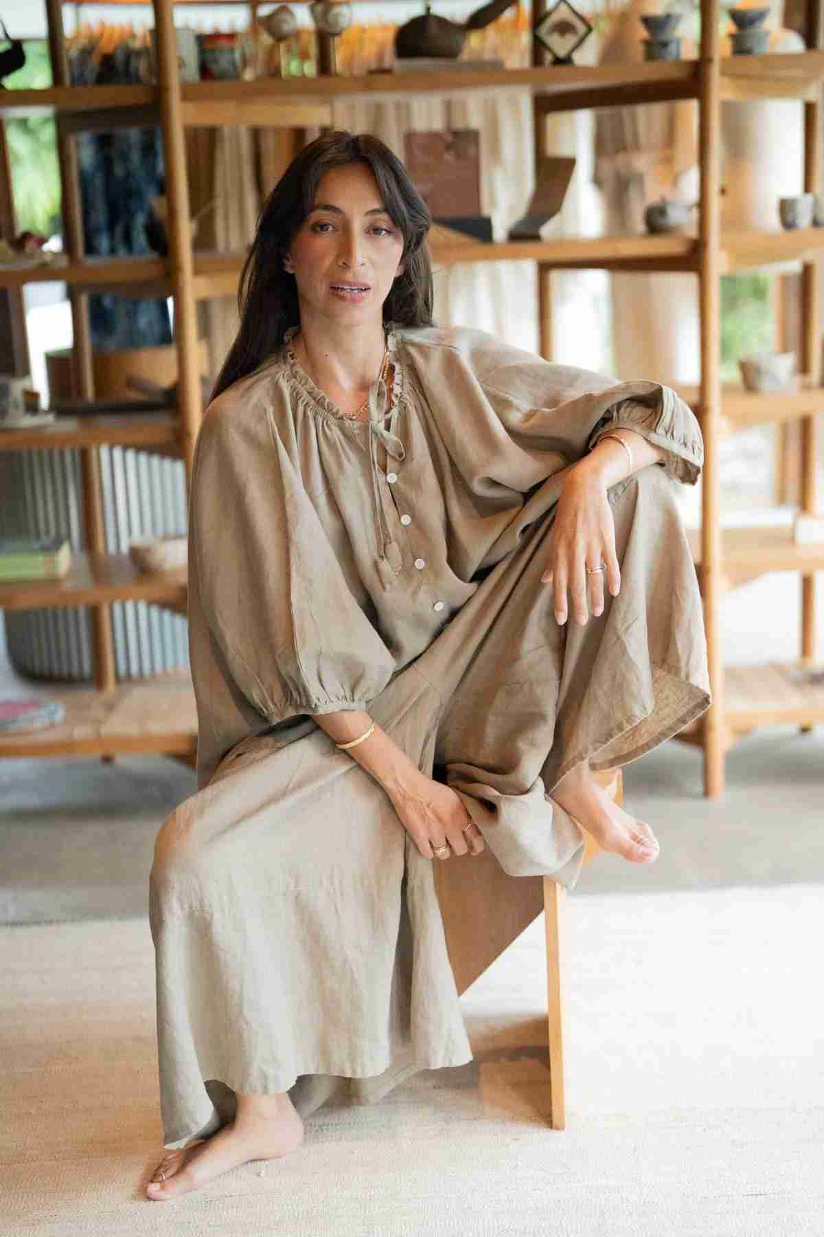 A person with long dark hair is seated on a chair in a relaxed pose, wearing an earthy-style outfit with a comfortable fit. They are surrounded by wooden shelves displaying various items in the background.