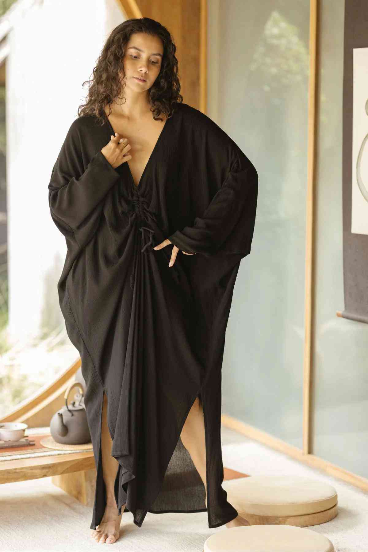 A woman stands indoors wearing a loose-fitting, flowing black Bamboo Rayon Virgo Rising Gown by Myrah Penaloza. The dress boasts long sleeves and a deep V-neckline, with a cinched detail at the waist. The setting appears calm and minimalistic, complemented by a small table and tea set in the background.