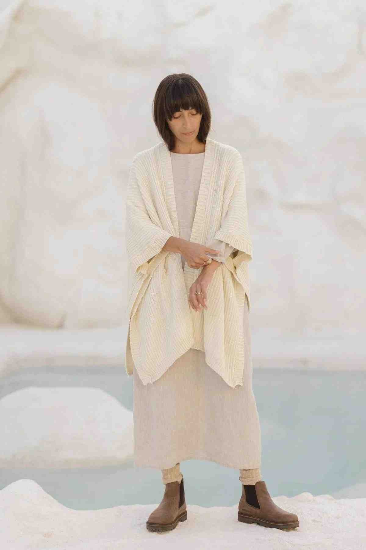 A person with shoulder-length dark hair stands with their arms crossed in front of a light-colored, rocky background. They are wearing the "Mountain Magic Linen Gown (Made To Order)" by Myrah Penaloza, paired with a long, off-white shawl and brown boots.