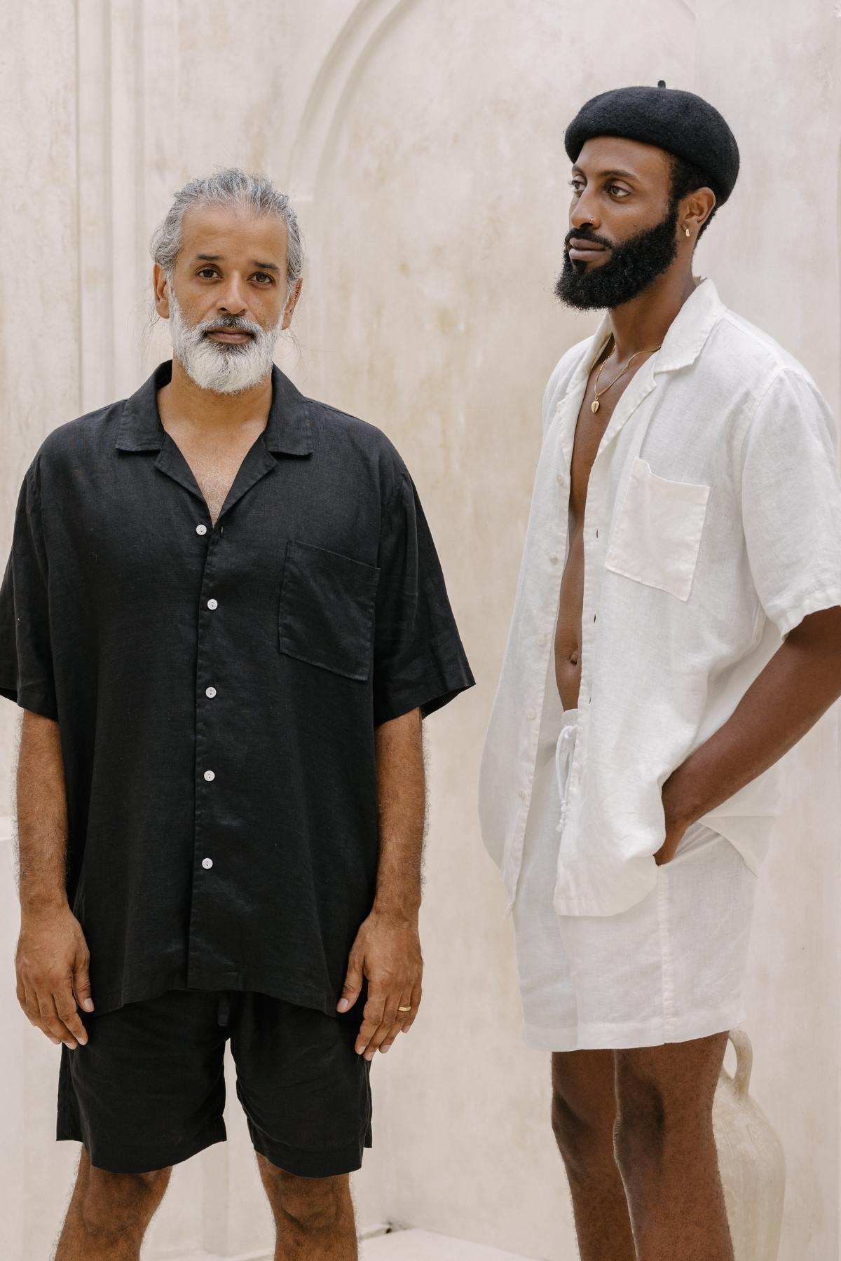 In a minimalist setting, two men stand with the relaxed expressions of nomadic travelers. The man on the left, with graying hair and a beard, wears a black Caravana Men's Linen Top & Short by Myrah Penaloza. Meanwhile, the man on the right dons a white Caravana Men's Linen Top & Short paired with a black beret. Their lightweight and breathable outfits emphasize their ease in movement.