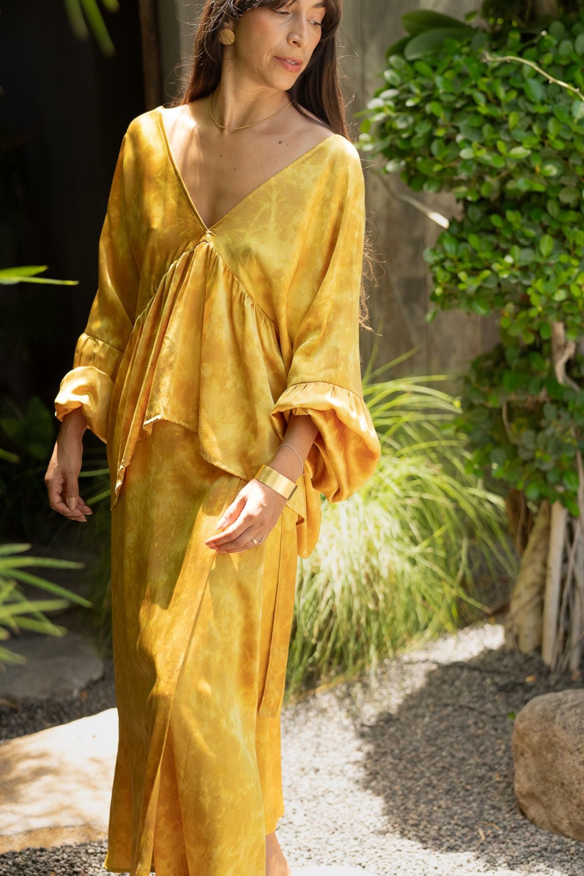 A person wearing the Eva Top & Shanti Skirt Set in a vibrant yellow with a floral design stands amidst lush greenery. Crafted from 100% naturally dyed silk, this made-to-order ensemble by Myrah Penaloza features flowing lines and long sleeves, with its environment-friendly fabric complementing the natural setting that resembles a garden or natural area.