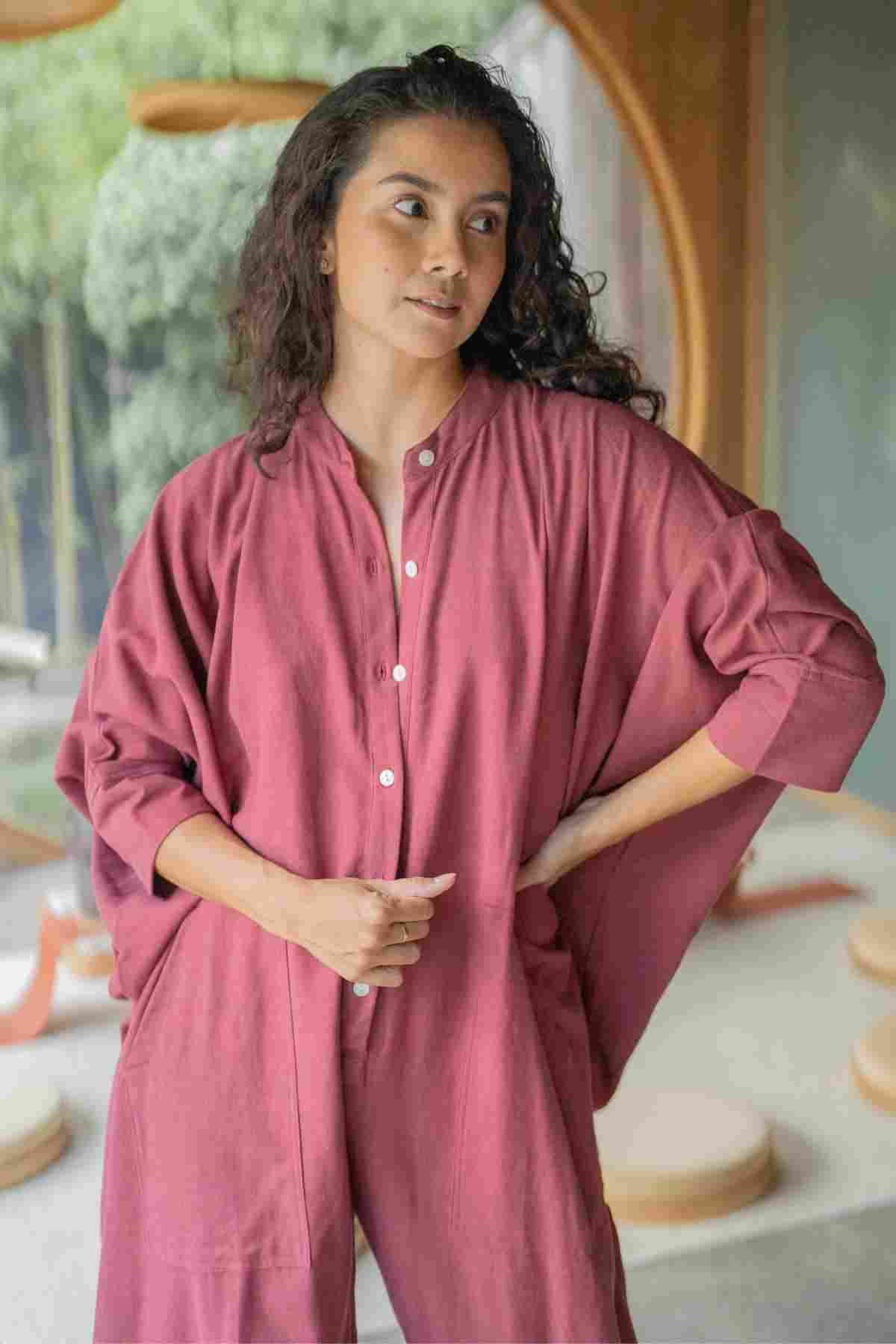 A person with long curly hair wears the Miracle Playsuit 2.0 (Karama Collection) from Myrah Penaloza and looks slightly to their left. They are standing indoors in a softly lit room with a large circular window and green foliage visible outside. Their hands rest lightly at their waist, embodying a boho chic vibe.