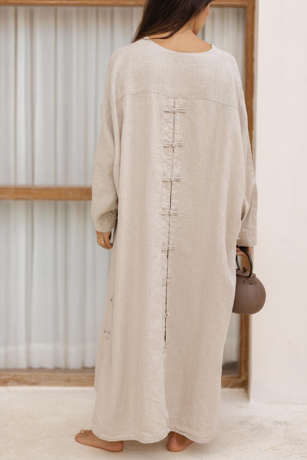 A woman stands barefoot facing away from the camera, wearing a loose, stone-washed Ancient Chajin Jacket (100% Linen) by Myrah Penaloza with a row of small Japanese-inspired buttons running down the back. She is holding a small brown handbag and standing in front of a wood-framed glass door with curtains.