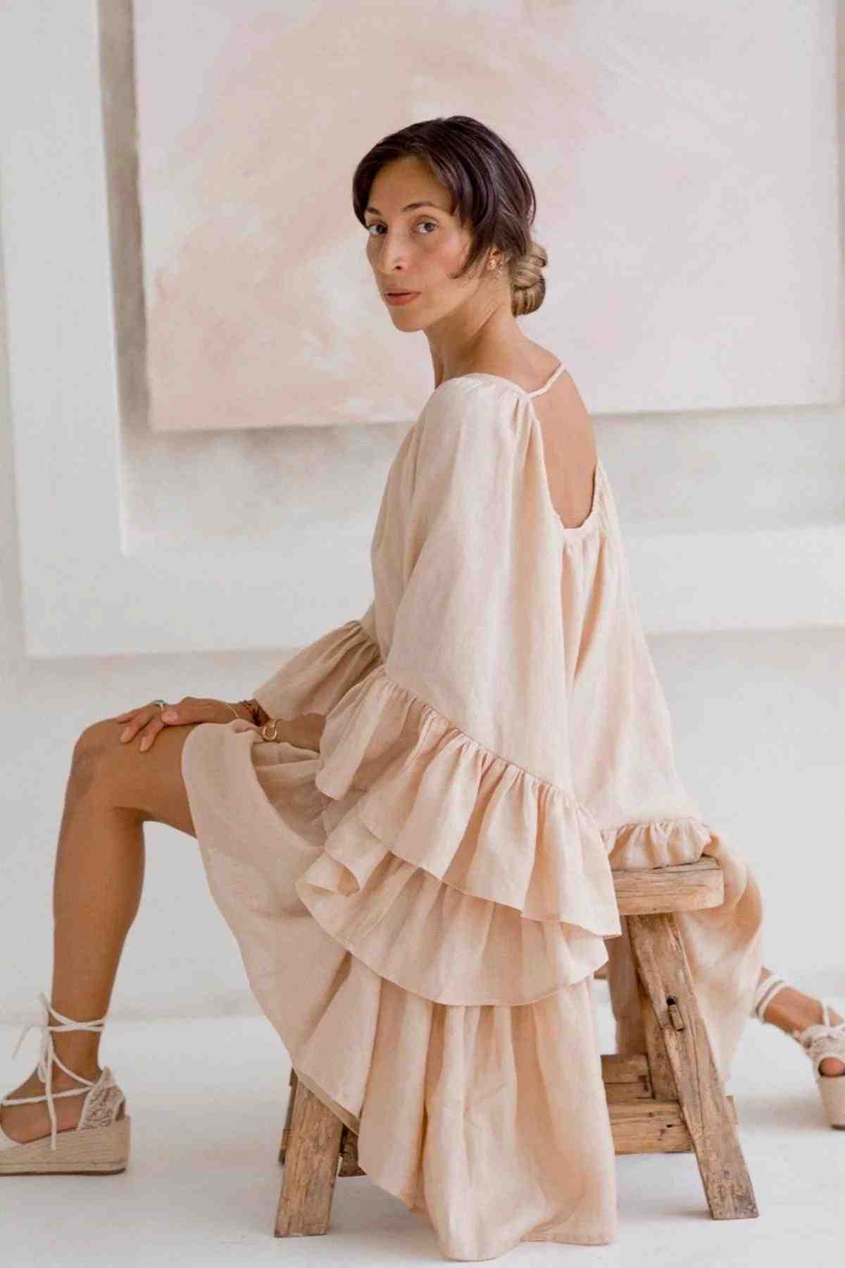 A woman with short brown hair sits gracefully on a wooden stool, wearing the Linen Brida Gown Short by Myrah Penaloza, perfect for spring and summer. The flowing cream gown features ruffled sleeves and is crafted from linen fabric. She looks over her shoulder against a soft, neutral background, complemented by her wedge sandals.