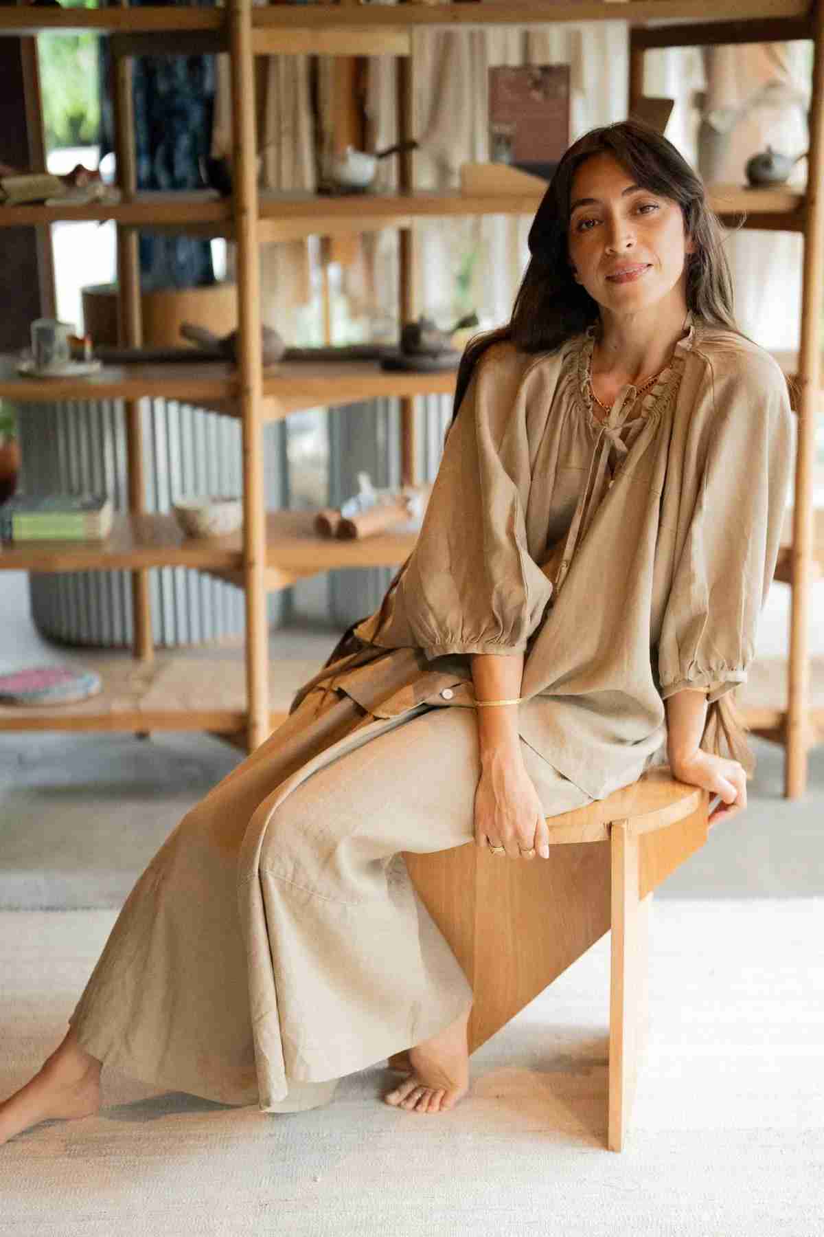 A woman wearing Myrah Penaloza's Pantalon Linen Pants, featuring a wide-legged design, sits on a wooden bench in a cozy room with wooden shelves and soft lighting. Her long dark hair and calm expression perfectly embody the casual and relaxed setting.