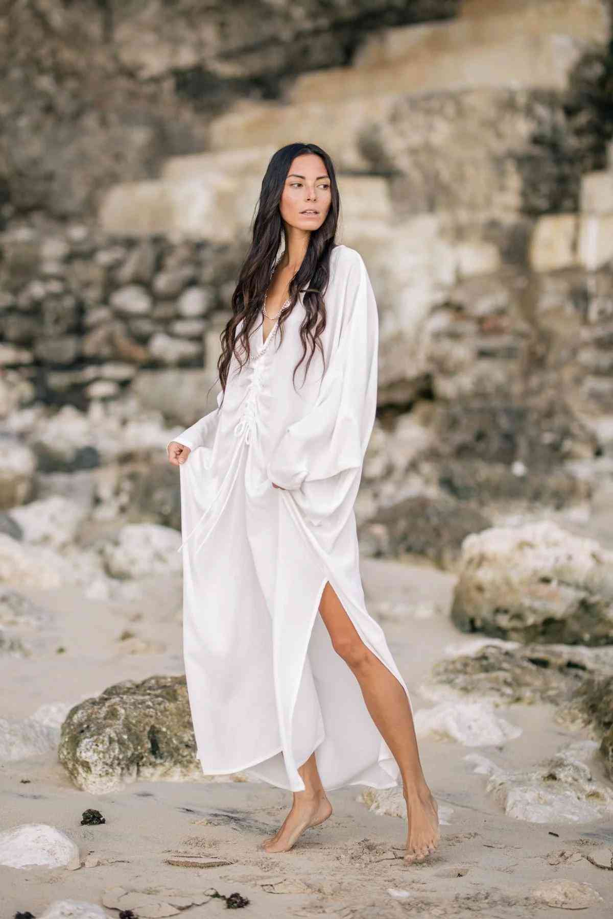A woman with long dark hair stands barefoot on a rocky beach, draped in the flowing Bamboo Rayon Virgo Rising Gown by Myrah Penaloza. She gazes into the distance, holding the sides of her dress. Large rocks and a cliffside form the background.