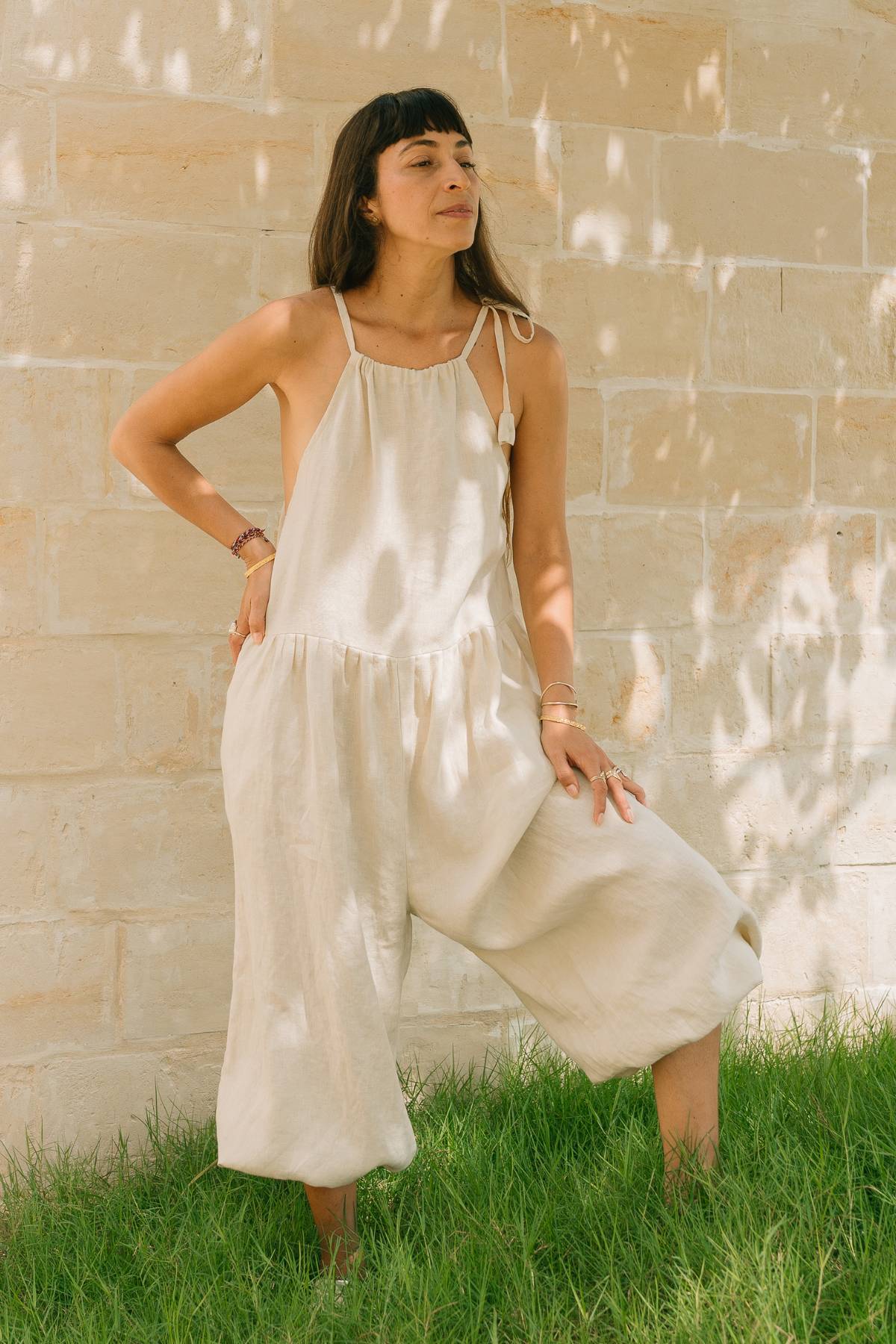 Woman wearing the Monaya Playsuit by Myrah Penaloza in Moonlight stone washed linen, one hand on hip, standing on grass against a stone wall. Oversized full length silhouette, handcrafted in Bali.