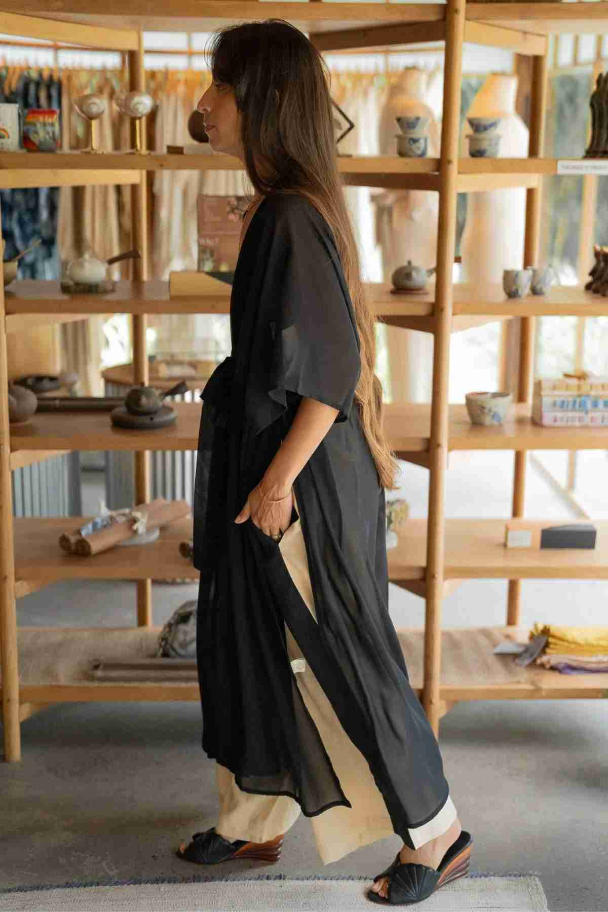 A person with long hair is standing sideways in front of a wooden shelf filled with various decorative items. They are wearing the Madelin Sheer Chiffon Long Cover by Myrah Penaloza over light-colored pants and black sandals, bringing sheer brilliance to the indoor, well-lit room.
