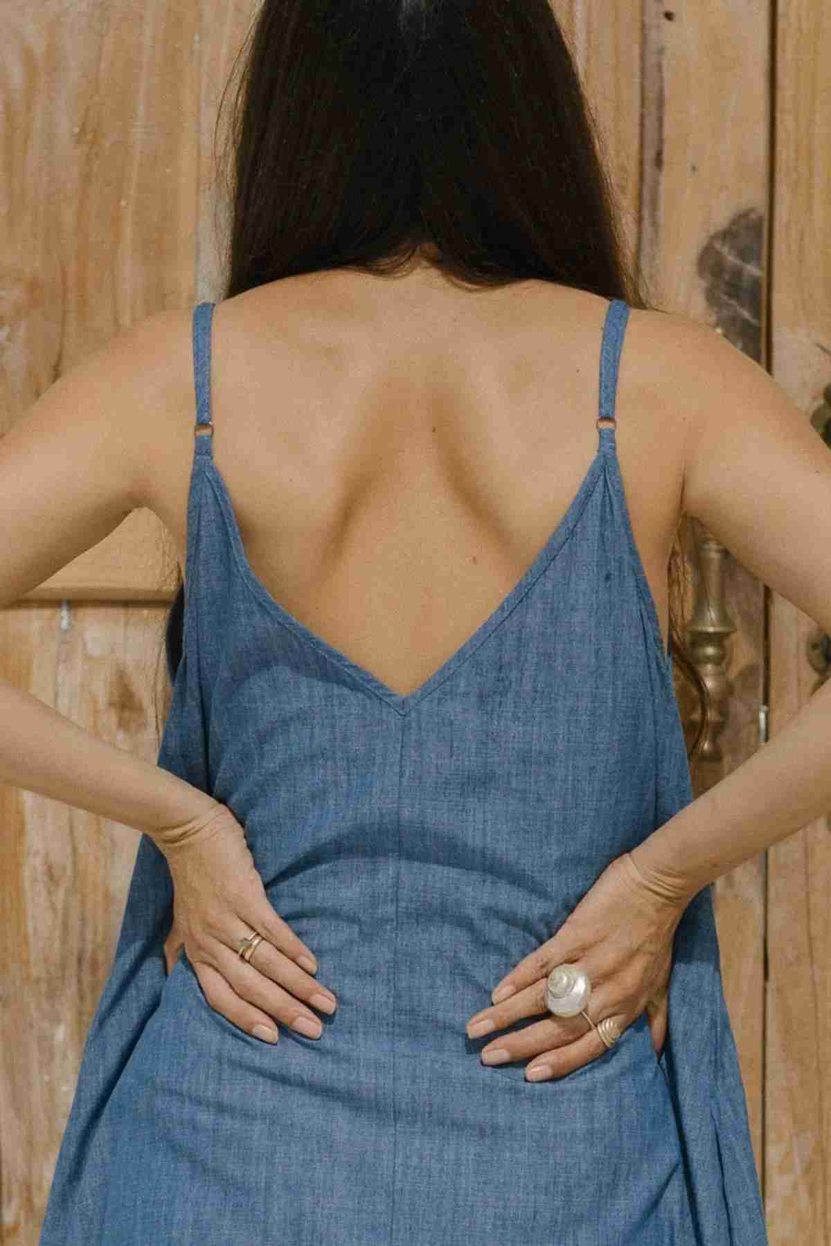 A person with long, dark hair is seen from behind, wearing the versatile Kundalini Denim Dress by Myrah Penaloza. They have their hands placed on their lower back, adorned with rings on multiple fingers. The background consists of a wooden door.