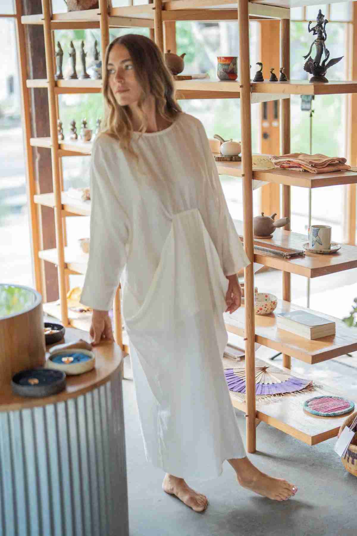 A woman in a long, flowing Mountain Magic Linen Gown by Myrah Penaloza stands barefoot in a room filled with wooden shelves displaying various items like pottery, figurines, and woven crafts. She is looking off to her right, with natural light illuminating the scene from large windows.