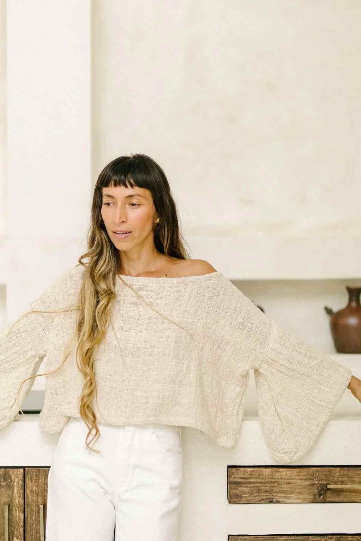 A person with long hair wearing an oversized, off-the-shoulder Tumanggal Cotton Spirit Top by Myrah Penaloza and white pants stands in a neutral-toned modern kitchen. Their arms are resting on the counter behind them, and they look serene and relaxed.