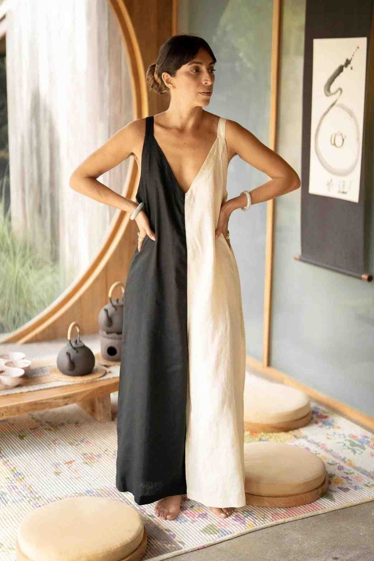A person wearing the Kundalini Linen Playsuit by Myrah Penaloza, featuring a bold, split black and white design, stands with their hands on their hips in a room with circular windows. A low table adorned with teapots and floor cushions surrounds them, while a scroll with calligraphy art hangs on the wall behind them, completing this luxuriously soft stonewashed linen setting.