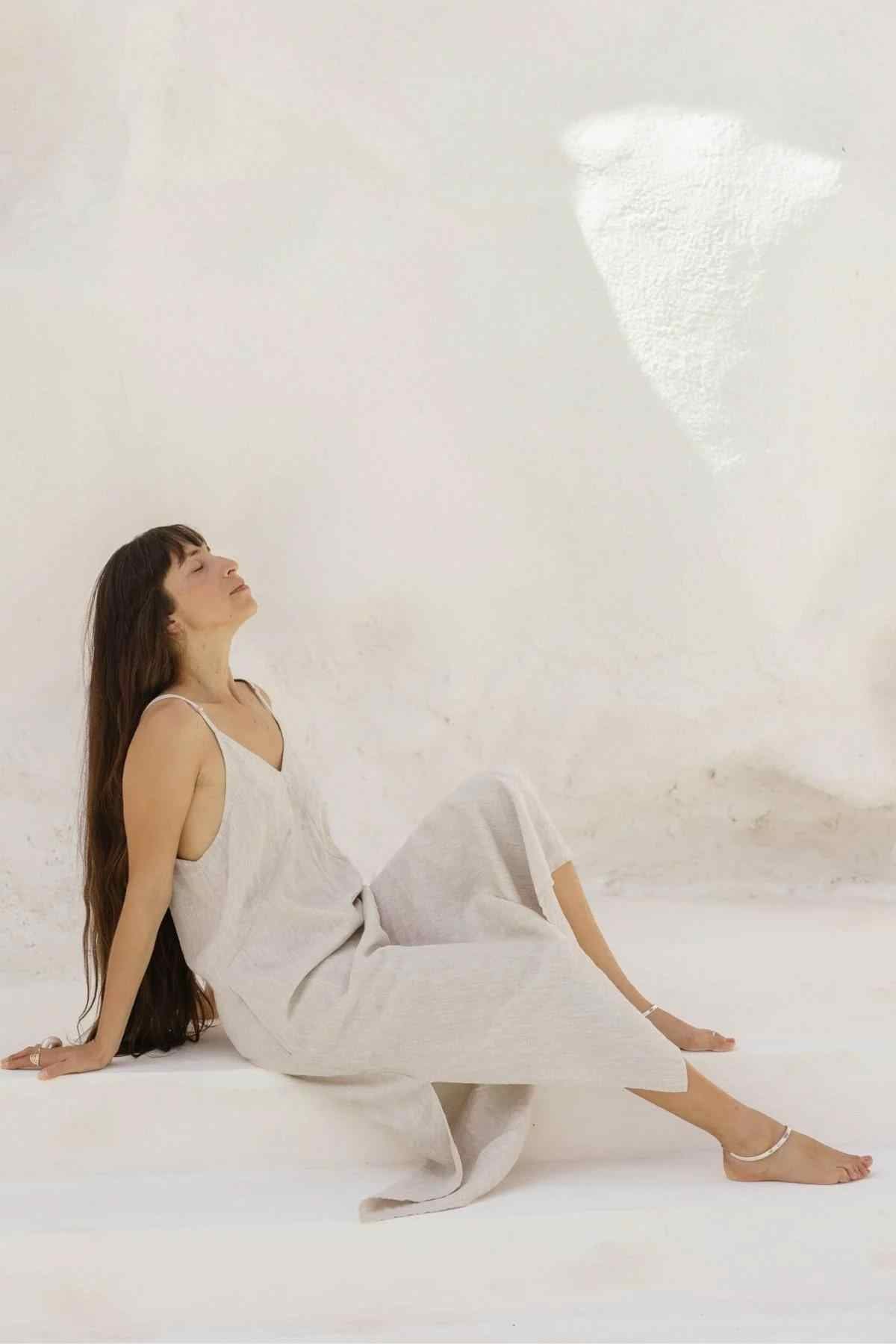 A woman with long dark hair sits on the ground against a light-colored textured wall, epitomizing luxurious comfort. She is wearing the Everyday Linen Slip by Myrah Penaloza, a sleeveless, ankle-length beige dress made of Stone Washed Linen, and white sandals. Her head is tilted back with her arms extended behind her to support her relaxed pose.