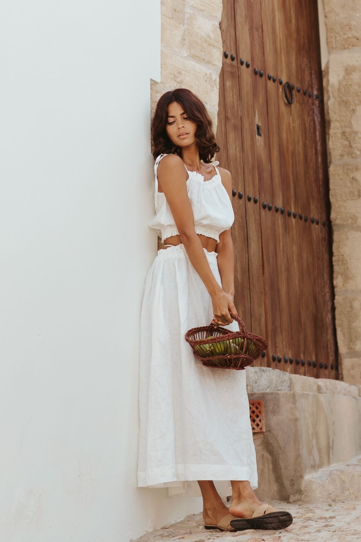 A woman wearing the Mona Top by Myrah Penaloza, characterized by its white sleeveless design and adjustable straps, leans against a stone wall close to a wooden door. She holds a wicker basket filled with green fruits while sporting brown sandals as she stands on the cobblestone street. Her dark hair cascades loosely as she looks downward in this timeless everyday scene.