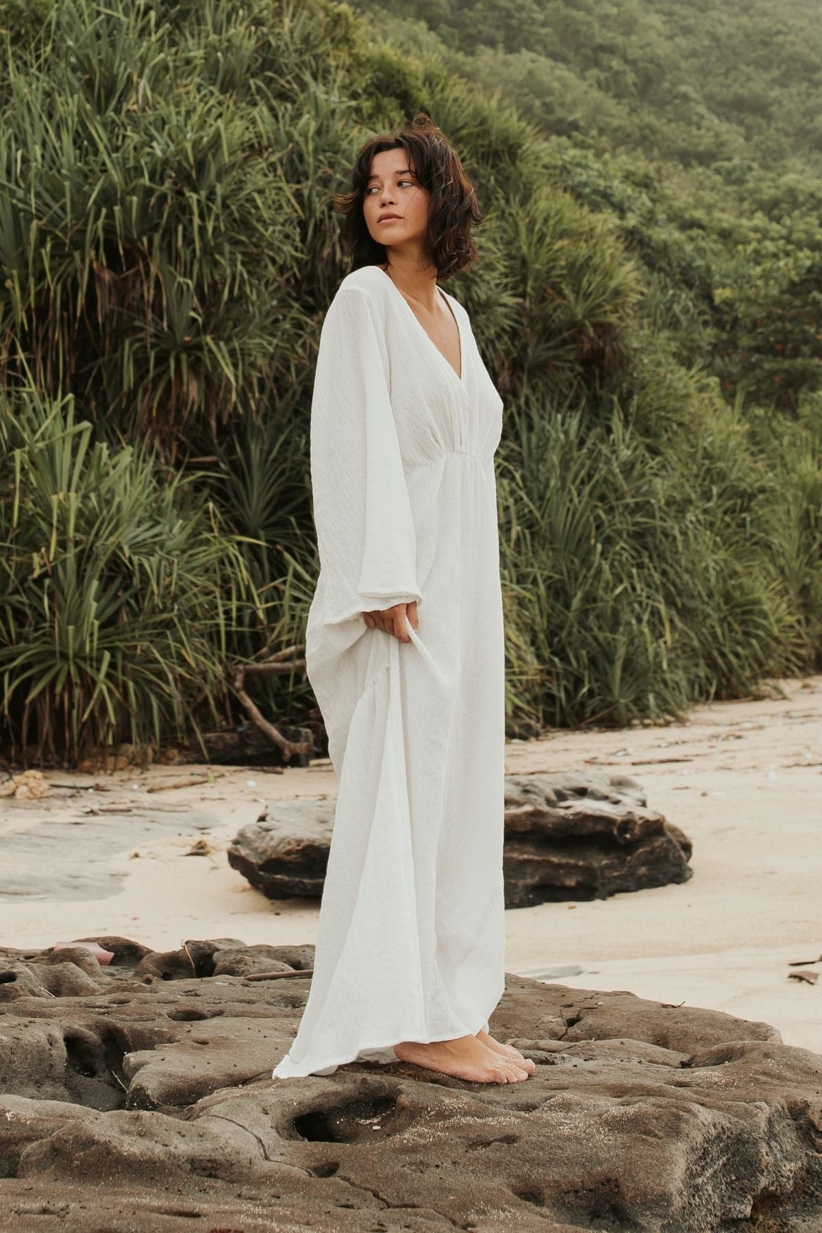 A person with shoulder-length, wavy hair stands on a rocky shore, wearing the elegant Sayan Gown (Linen/Cotton) from Myrah Penaloza. The gown features an open back and long sleeves. They are barefoot and surrounded by lush green foliage and sand, gazing off to the side to create a serene, reflective atmosphere.