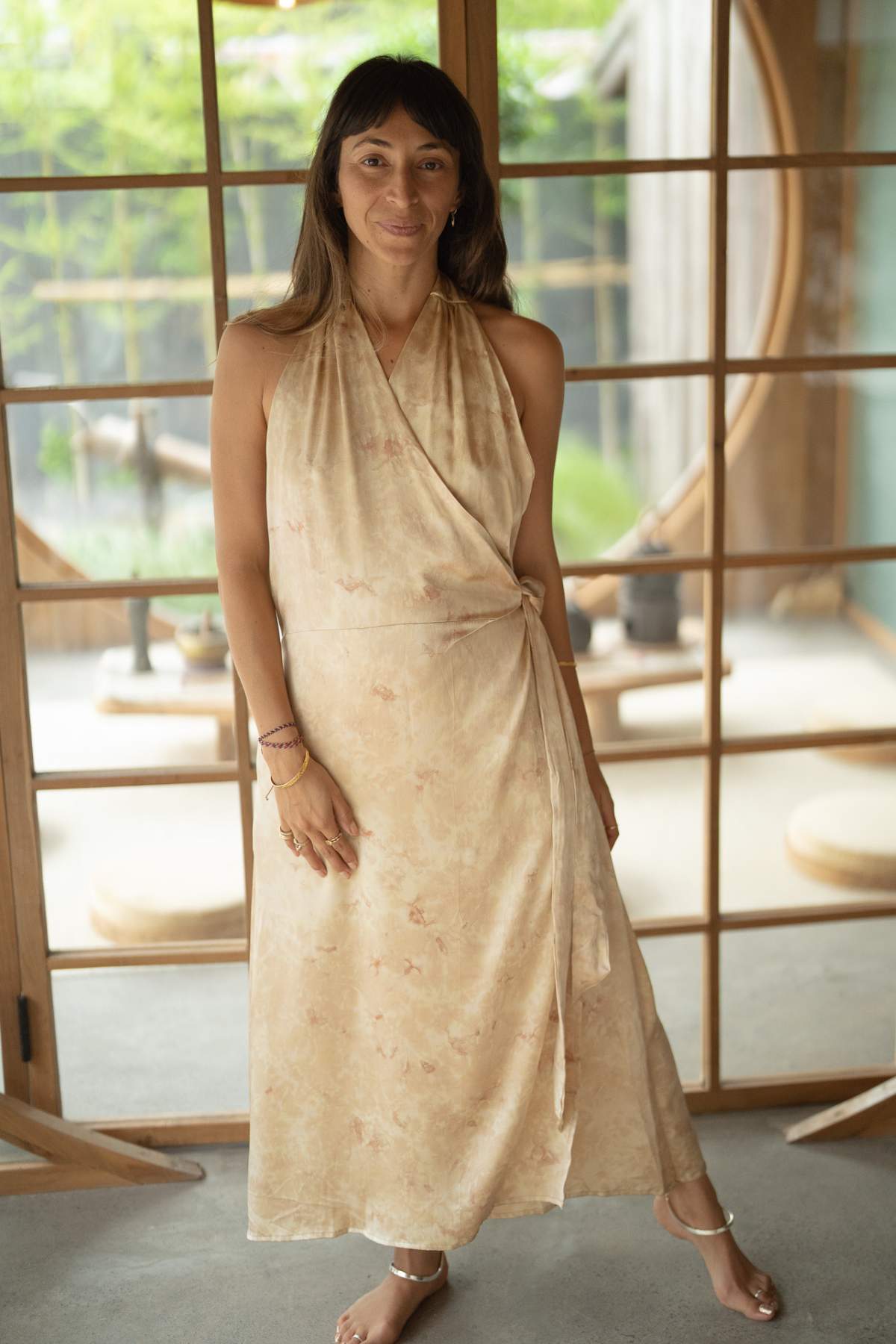 In a serene indoor setting, a woman exudes elegance in the Ratu Silk Wrap Gown by Myrah Penaloza. The light beige dress, botanically hand-dyed on silk with subtle patterns, beautifully complements her flowing hair. She smiles serenely, framed by a wooden sliding door and lush greenery that serves as her regal backdrop.