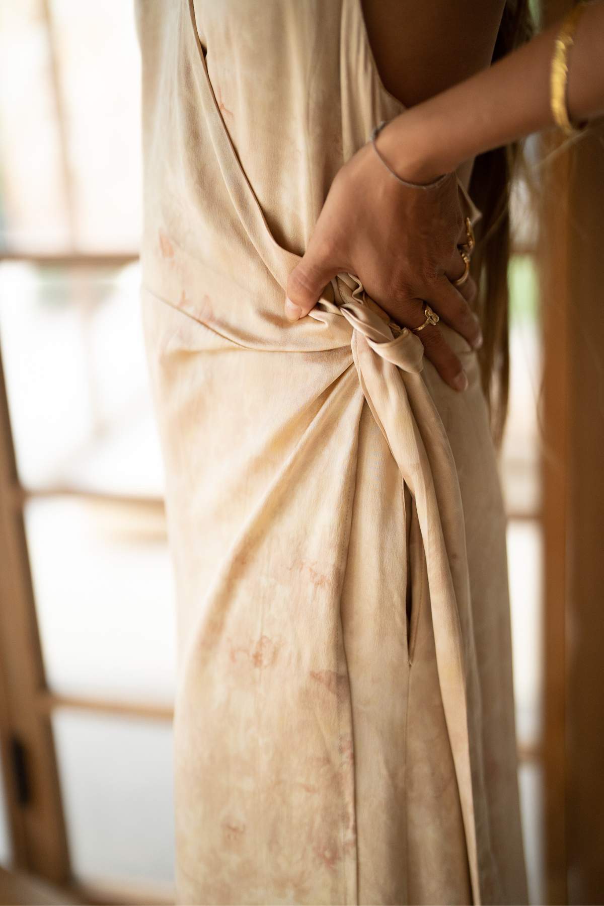 Standing near a window with wooden frames, a person adjusts the tie on the side of their Ratu Silk Wrap Gown by Myrah Penaloza. The botanically hand-dyed soft beige dress boasts a flowing, draped design, while delicate rings and a bracelet adorn the person's hands.