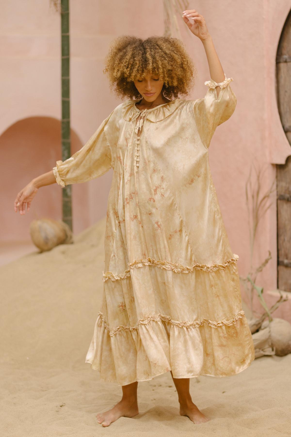 A person with curly hair is wearing the Allegra Joy, a hand-dyed rose petal silk gown with tiered ruffles by Myrah Penaloza. They are standing on sandy terrain with one arm raised and the other slightly bent. The background includes arched doorways and a rustic, earthy setting, evoking a sense of vintage couture.
