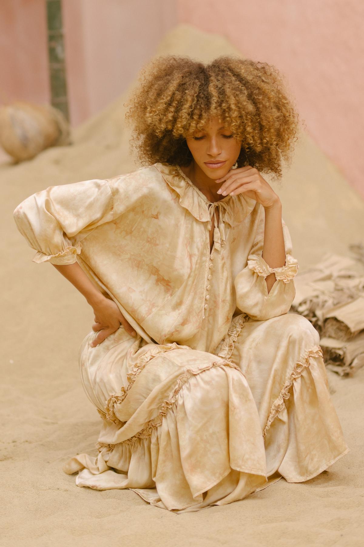 In a minimalist, earthy-colored setting framed by pale pink walls and dry elements, a woman with curly hair sits pensively on the sandy ground. She is clad in the Allegra Joy, Rose Petal Dyed, Silk Gown - Made To Order by Myrah Penaloza. The flowing beige gown features delicate ruffles, and as she rests her chin on her hand with a calm expression, the vintage couture scene comes to life.