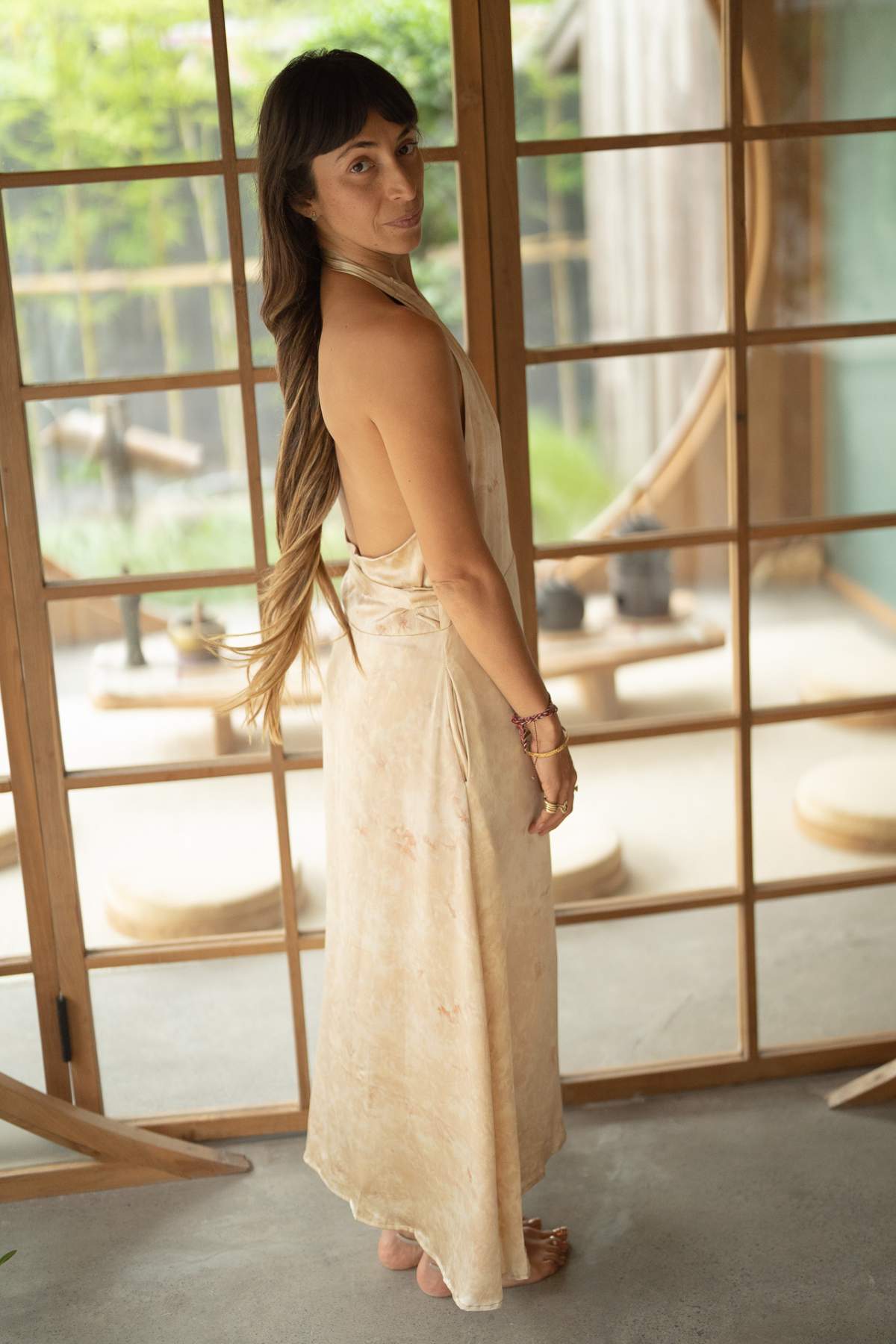 A woman with long brown hair stands in a room featuring wooden sliding doors and soft lighting. She is gracefully adorned in the Ratu Silk Wrap Gown by Myrah Penaloza, evoking the elegance of a queen as she glances over her shoulder. The background reveals a serene view of greenery outside.