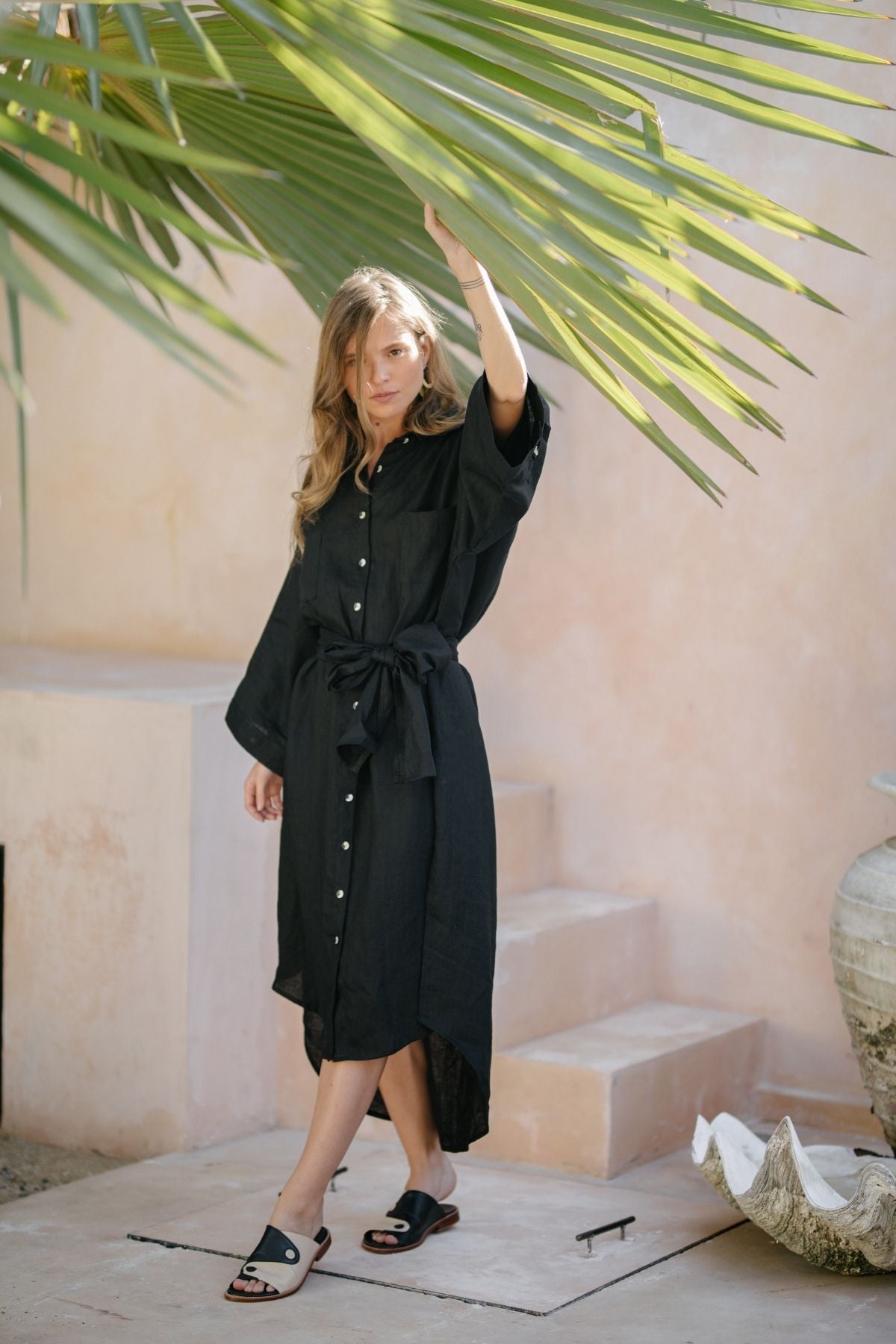 A woman wearing the Nidra Full Length Linen Duster by Myrah Penaloza stands on stone steps, partially shaded by large palm leaves. She holds one leaf with her hand while wearing black sandals against a peach-colored wall backdrop. An oversized pot is to her right.