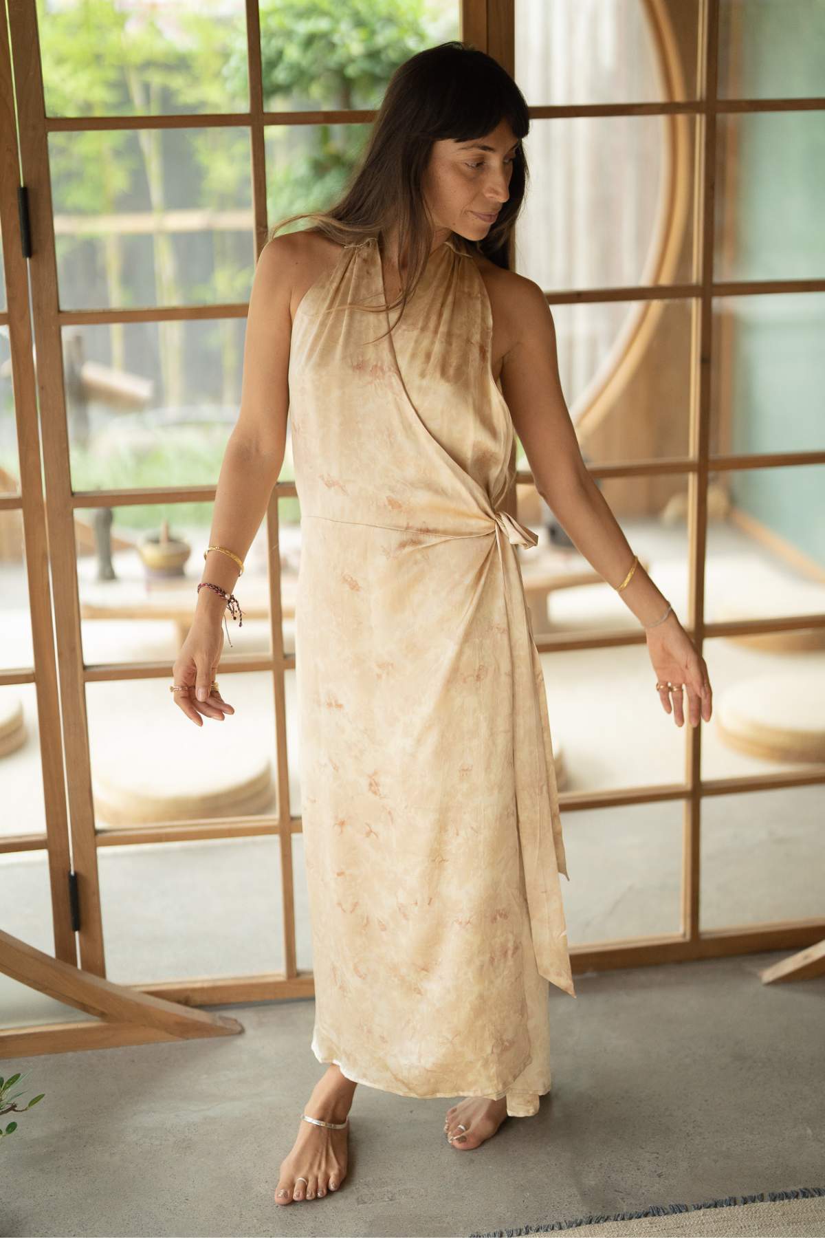 In a room featuring wooden sliding doors and a circular window, a woman stands elegantly in the Ratu Silk Wrap Gown by Myrah Penaloza. This sleeveless beige dress is botanically hand-dyed on luxurious silk, making her appear like a queen at rest. She gazes downward with her arms softly extended, adorned with minimal jewelry and wearing sandals.