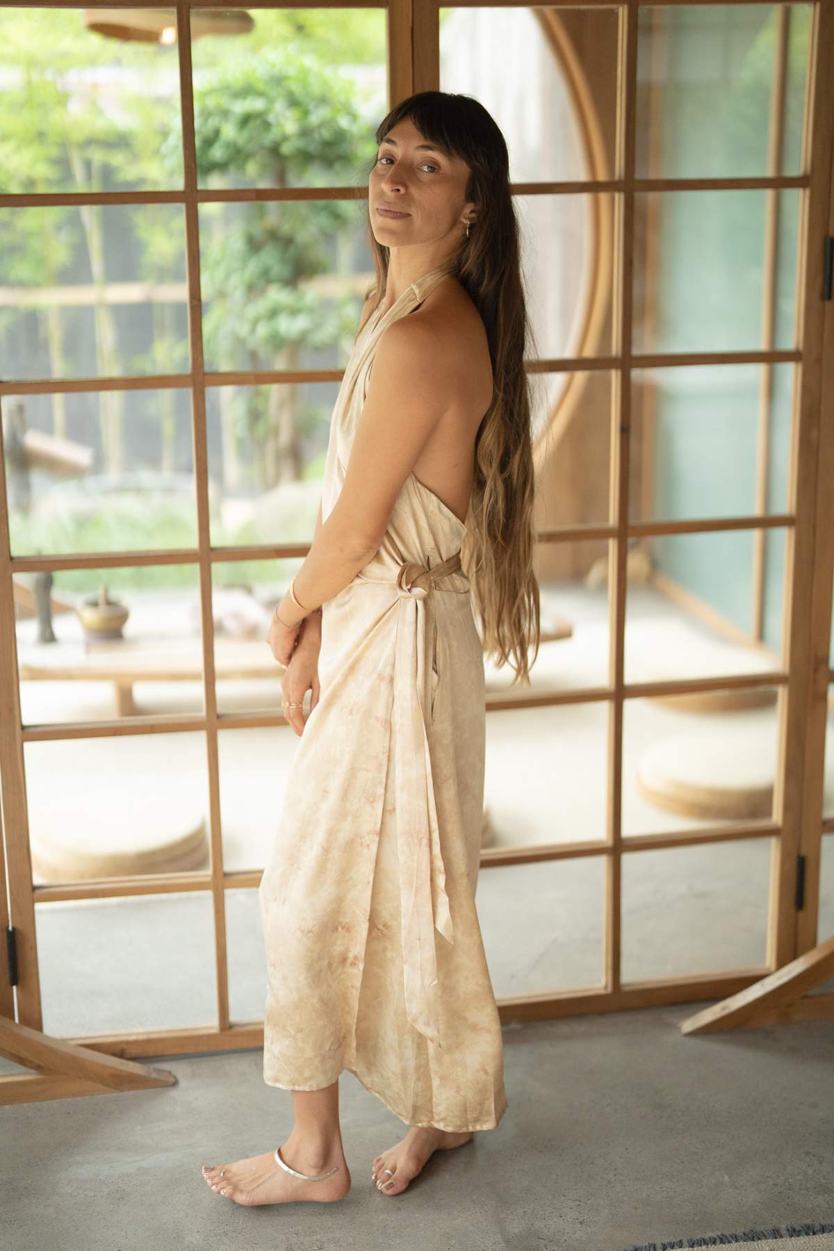 A woman with long hair stands indoors, elegantly dressed in the Ratu Silk Wrap Gown from Myrah Penaloza and sandals. She is turned slightly to the side, smiling softly like a queen, with wooden sliding doors and lush garden greenery visible in the background.