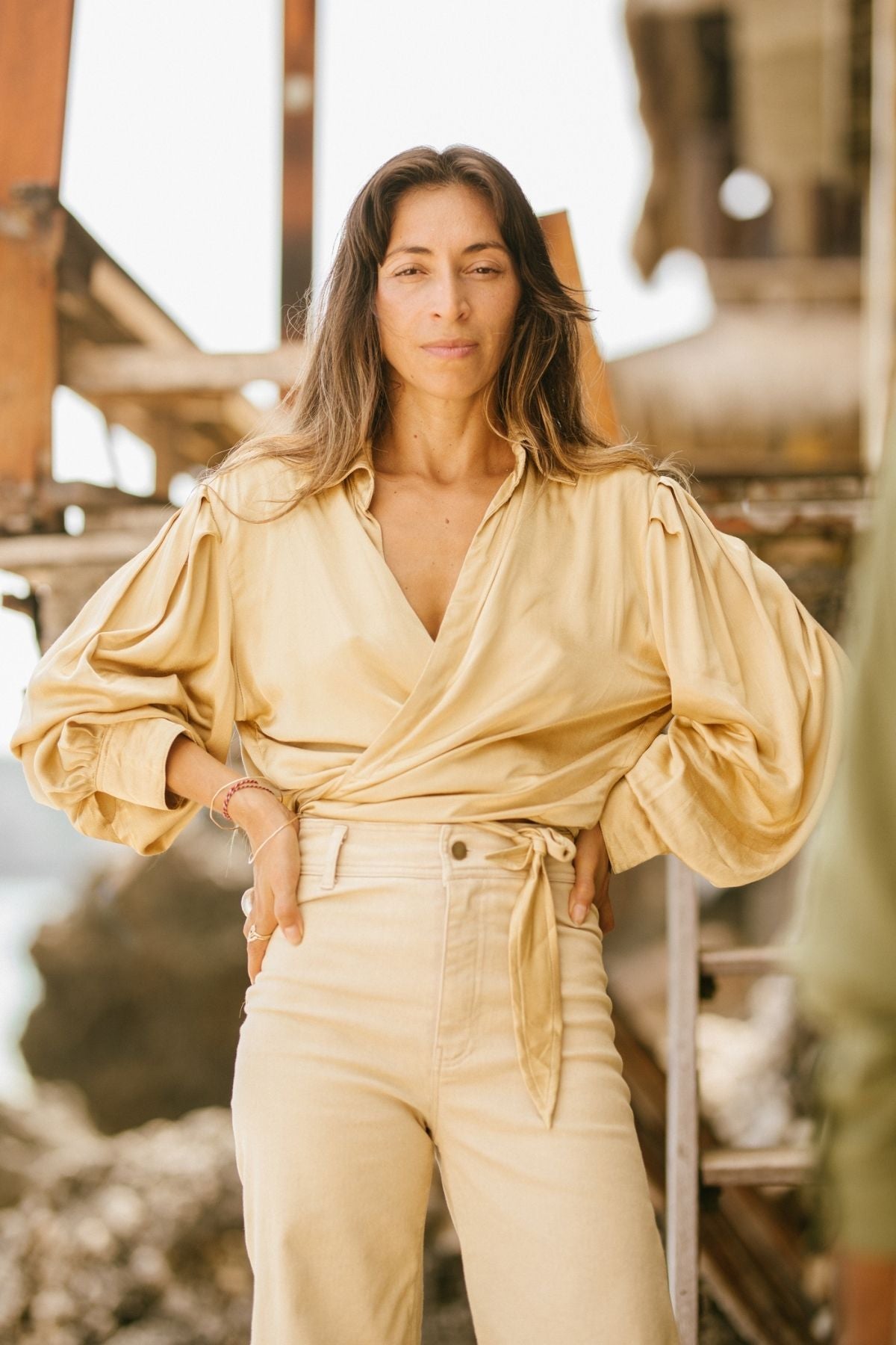 A woman stands confidently outdoors, dressed in a beige wrap blouse with puff sleeves and high-waisted Vida jeans from Myrah Penaloza. The stonewashed denim, botanically hand-dyed, accentuates her form perfectly. She has long, wavy hair and a neutral expression, with one hand resting on her hip. The background appears to be a rustic wooden structure.