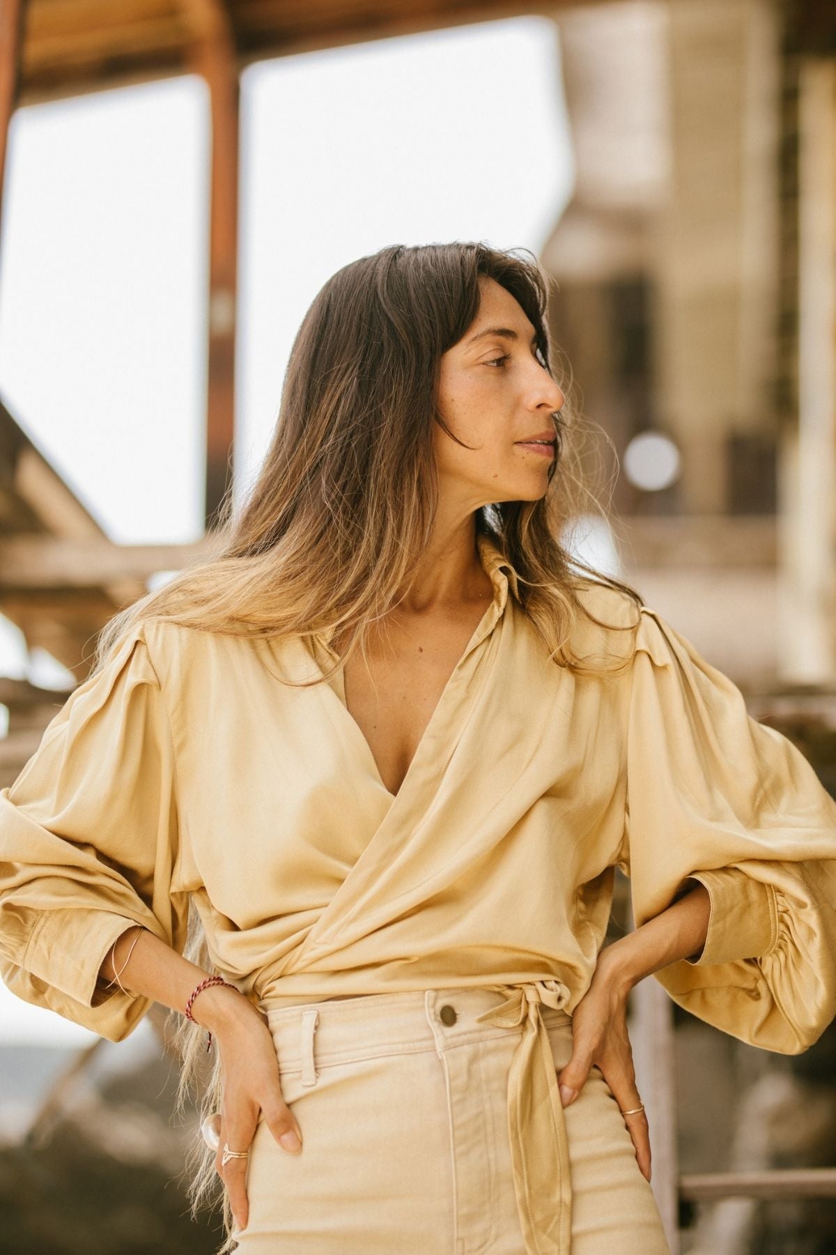 A person with long, wavy hair stands outside, wearing a light beige wrap-top paired with the Vida high-waisted jeans by Myrah Penaloza. These stonewashed jeans are botanically hand-dyed and made from stretchy denim. The person is positioned facing sideways with hands on their hips, against a blurred background of industrial-style architecture.