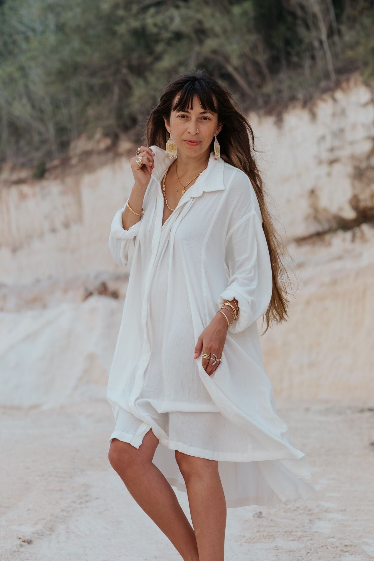 A woman with long hair walks confidently outdoors, wearing a flowing off-white Kundalini Gown Short by Myrah Penaloza, crafted entirely from bamboo rayon. Her statement earrings and rings complement the rocky, natural landscape beautifully.