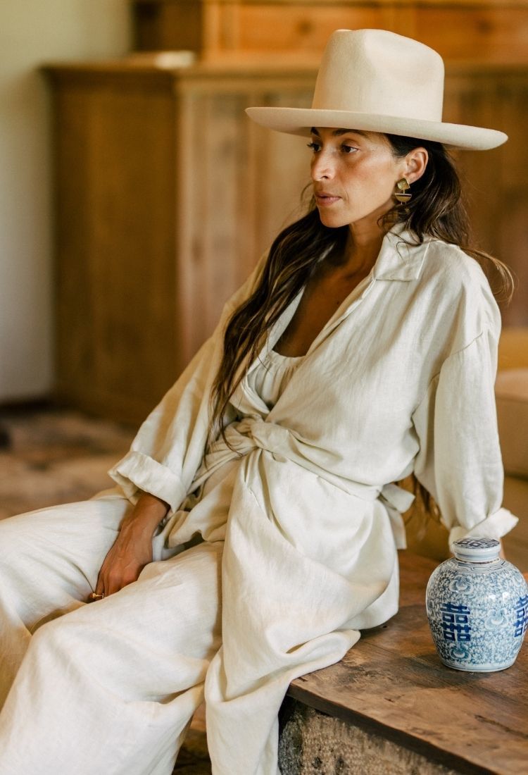 A person wearing the Infinity Jacket by Myrah Penaloza, crafted from 100% linen, sits on a wooden surface. They hold a decorative vase with blue patterns, set against an indoor background featuring soft lighting and a wooden backdrop.