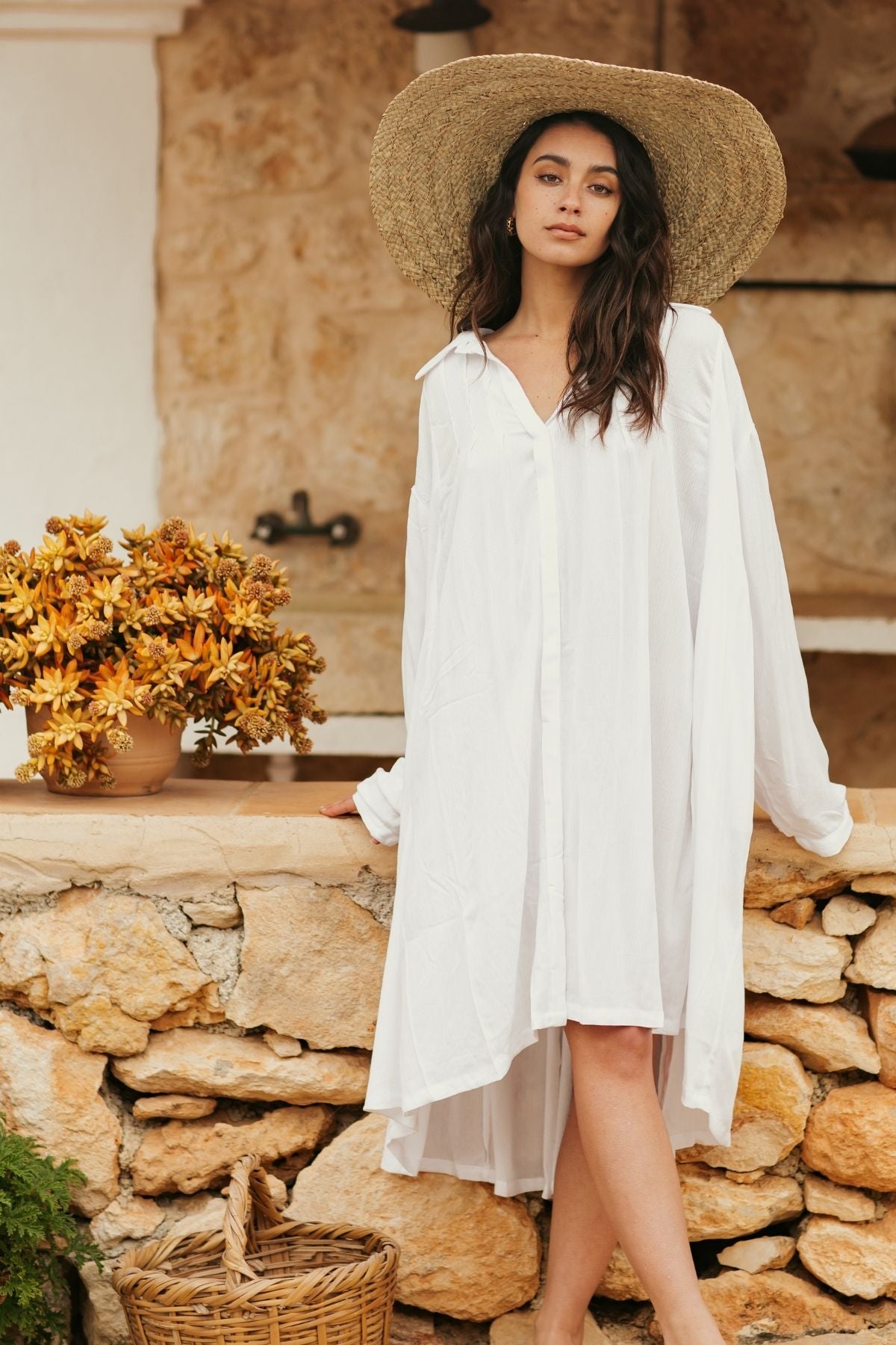 A woman wearing a Myrah Penaloza Kundalini Gown Short, made from 100% Bamboo Rayon in an off-white shade, and a wide-brimmed straw hat stands by a stone wall. Beside her, there's a basket and a pot filled with yellow flowers. The rustic stone building in the background enhances the elegance of her handmade garment.