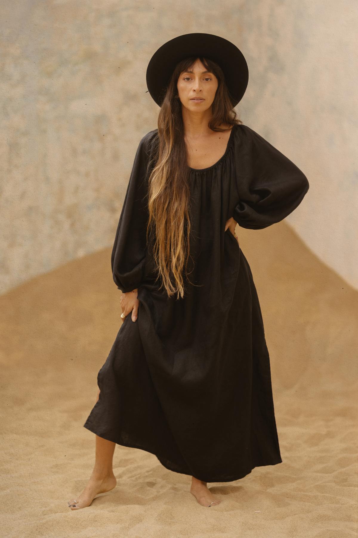 A modern mystic with long hair stands barefoot on sand, clad in a loose-fitting black Alchemy Gown (Crinkle Linen Cotton) by Myrah Penaloza and a wide-brimmed black hat. One hand rests on their hip while the other lightly holds the versatile gown. The neutral, textured backdrop enhances the earthy and serene setting.