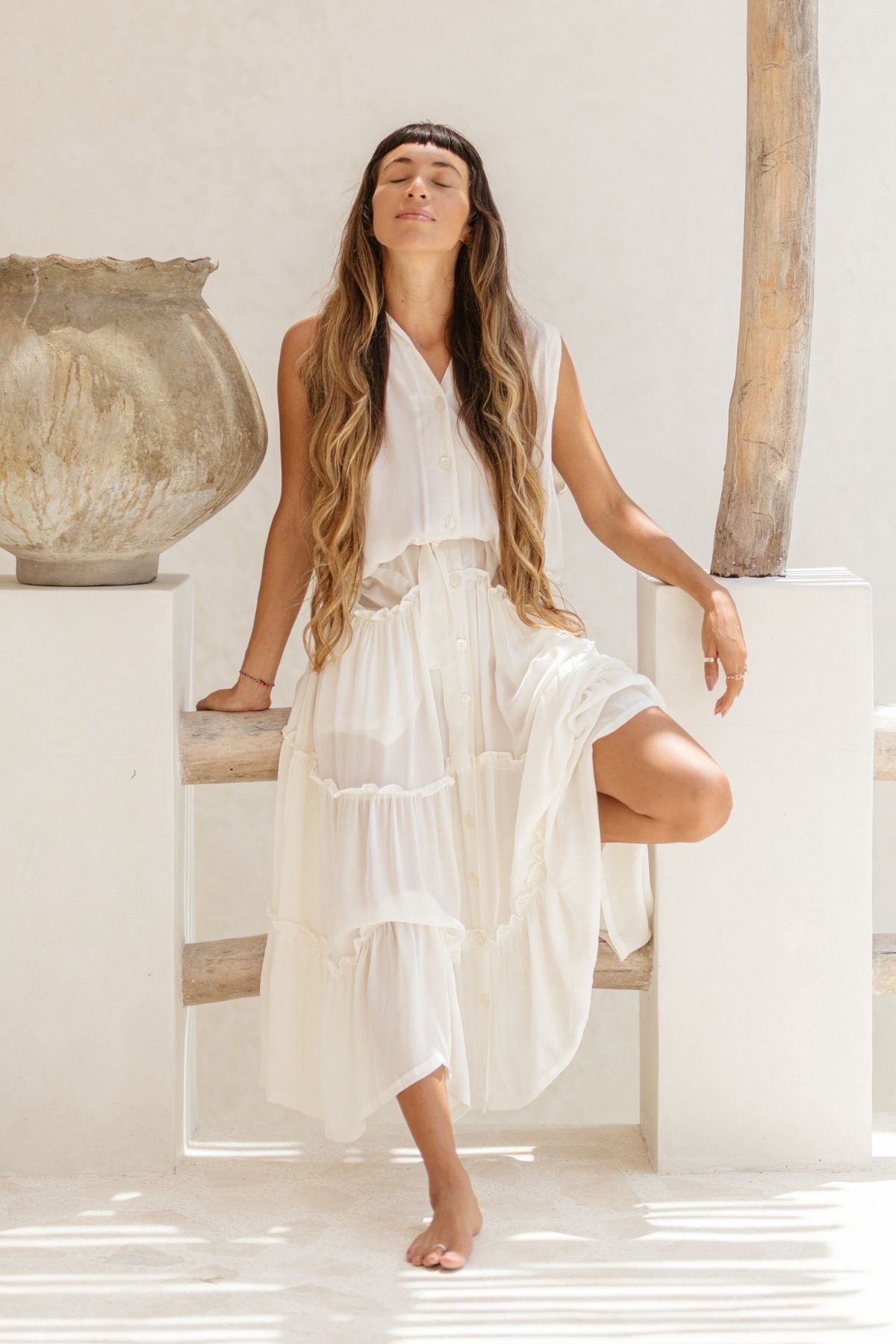 A woman in a sleeveless, ruffled-neck, 100% silk gown from Myrah Penaloza's La Dama collection stands with one leg raised on a low wooden fence. Her eyes are closed and her head tilted back, while her long hair cascades down her shoulders. The scene is reminiscent of a renaissance-inspired setting and features a large rustic vase on a stone pedestal.