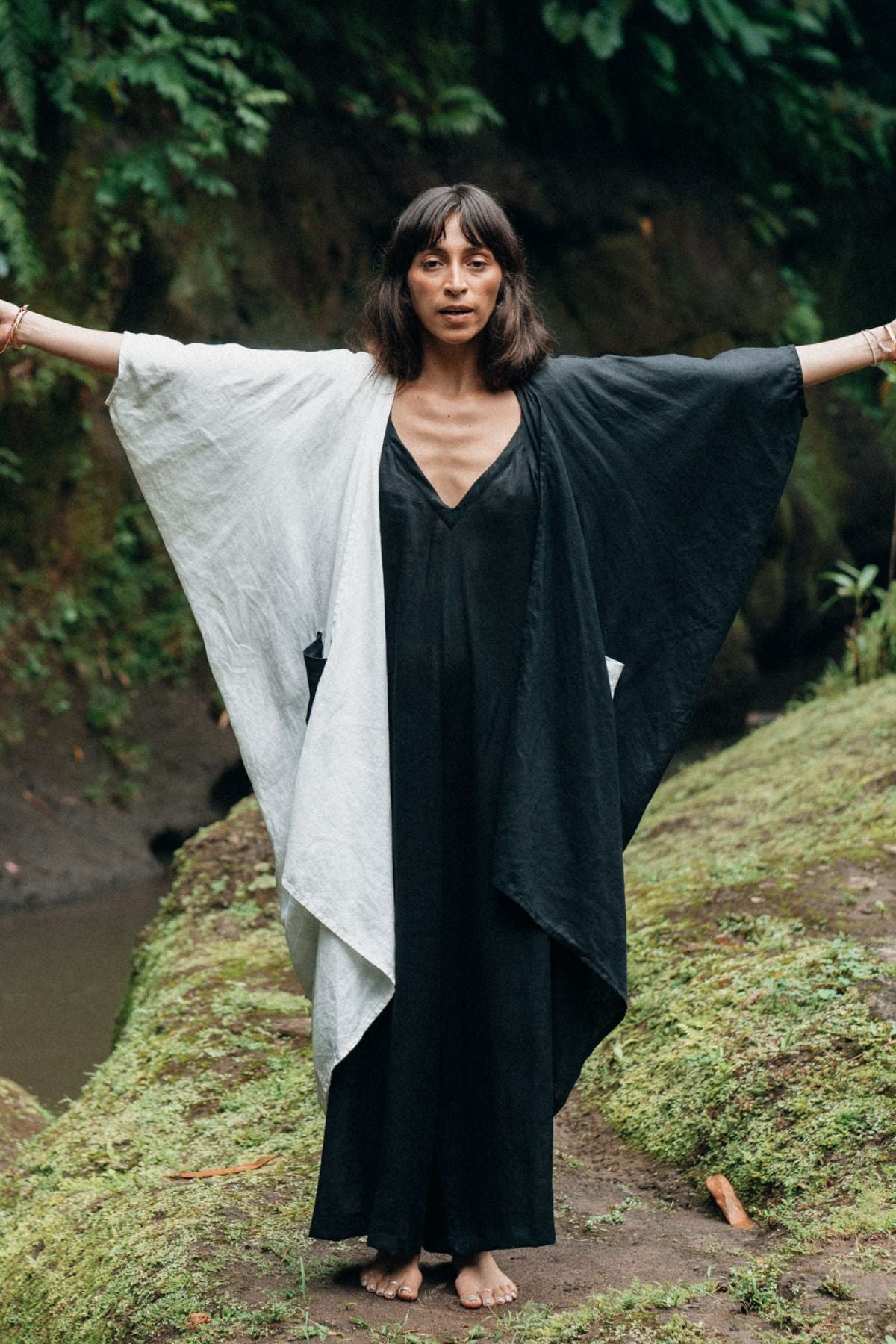 A woman stands outdoors on a mossy path with her arms outstretched, draped in a bicolor shawl over her flowing Dao Robe by Myrah Penaloza. The backdrop of lush greenery and a small stream enhances the tranquil scene.