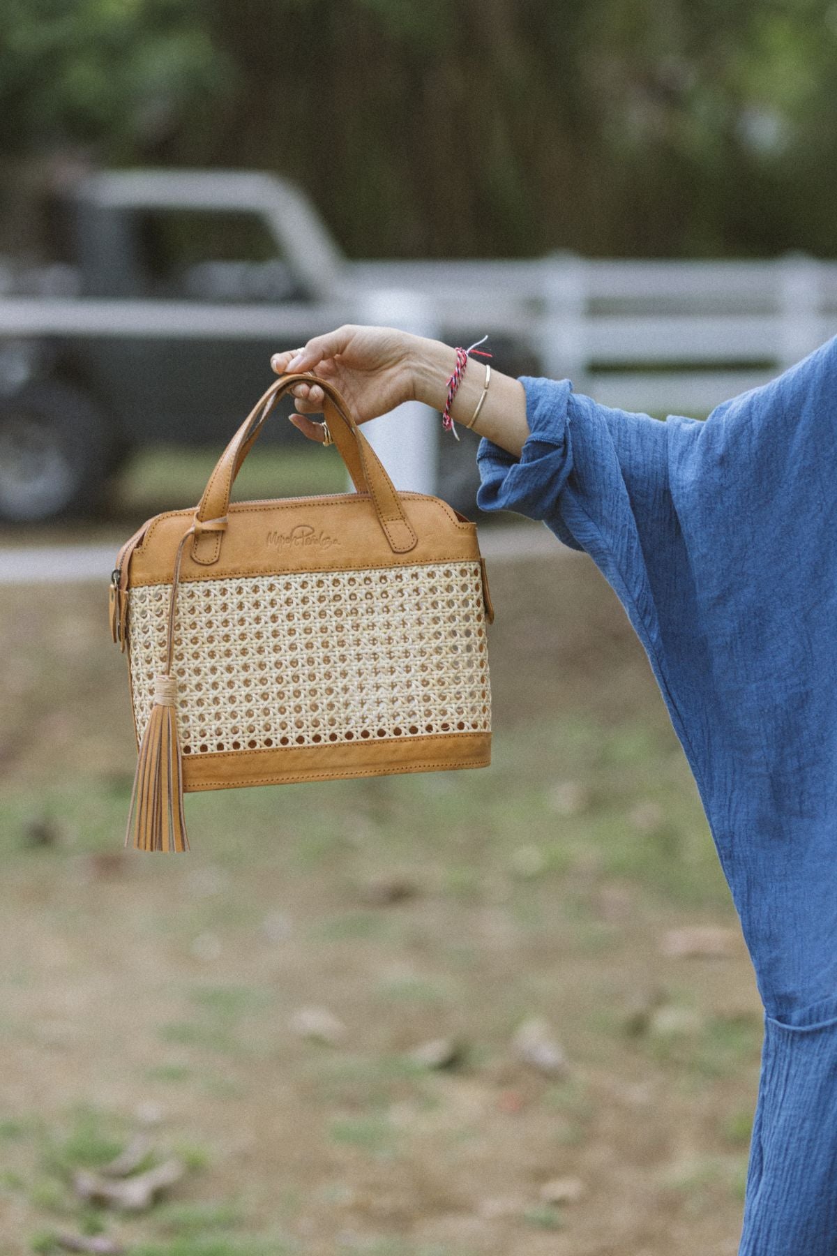 A person wearing a blue sleeve holds up a stylish Wonder Everyday Tote Mini by Myrah Penaloza, featuring a handcrafted Bali design with a woven rattan pattern and tan leather accents, including a tassel, set against an outdoor backdrop with a blurry vehicle and fence in the background.