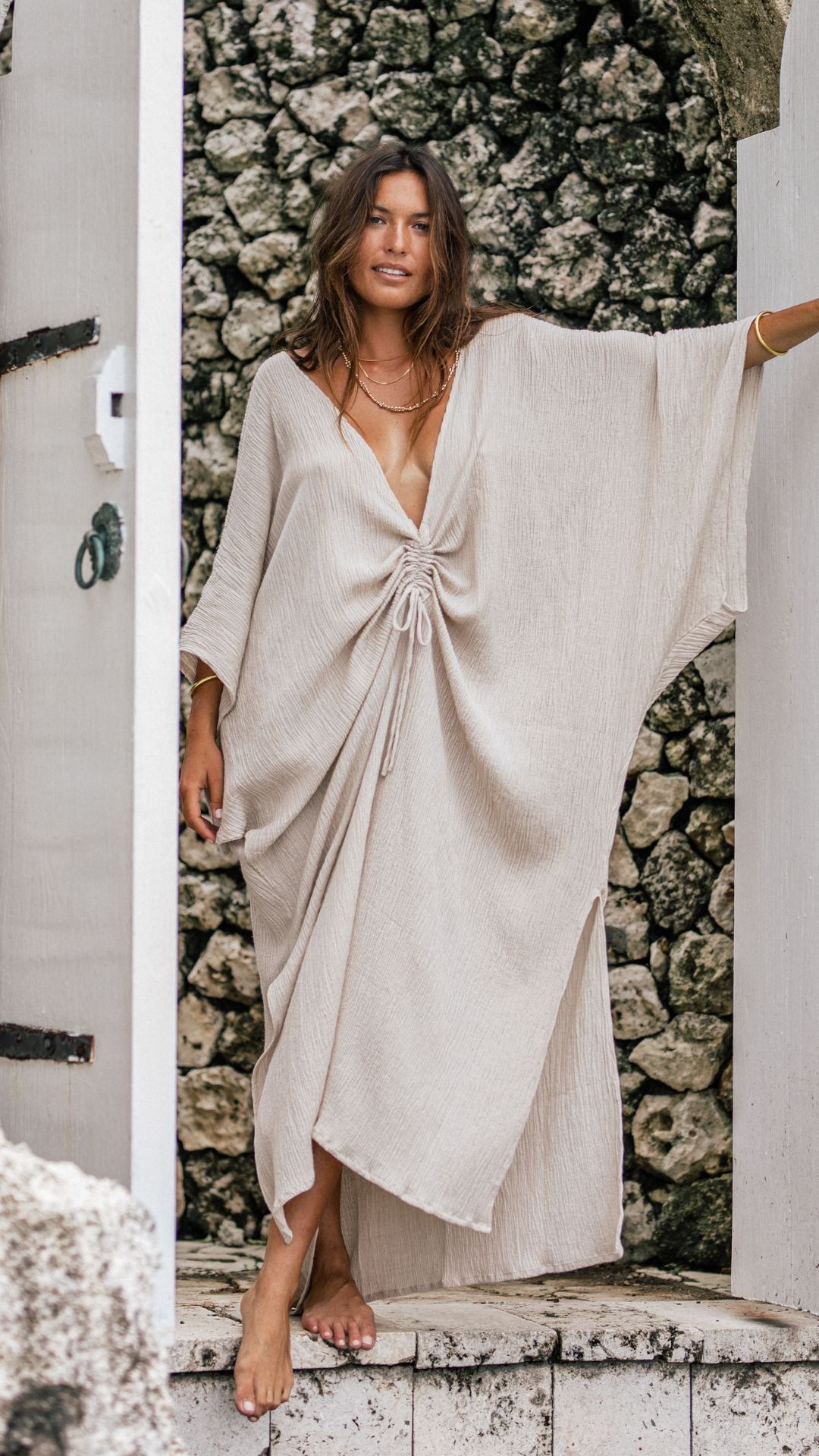 A woman stands barefoot in a doorway with a stone wall behind her, embodying natural beauty in the Limited Edition Virgo Kaftan by Myrah Penaloza. The light-colored garment, crafted from 100% crinkle linen/cotton in a natural hue, features a deep neckline and three-quarter sleeves, tied gracefully at the waist, as her hair cascades loosely around her shoulders.