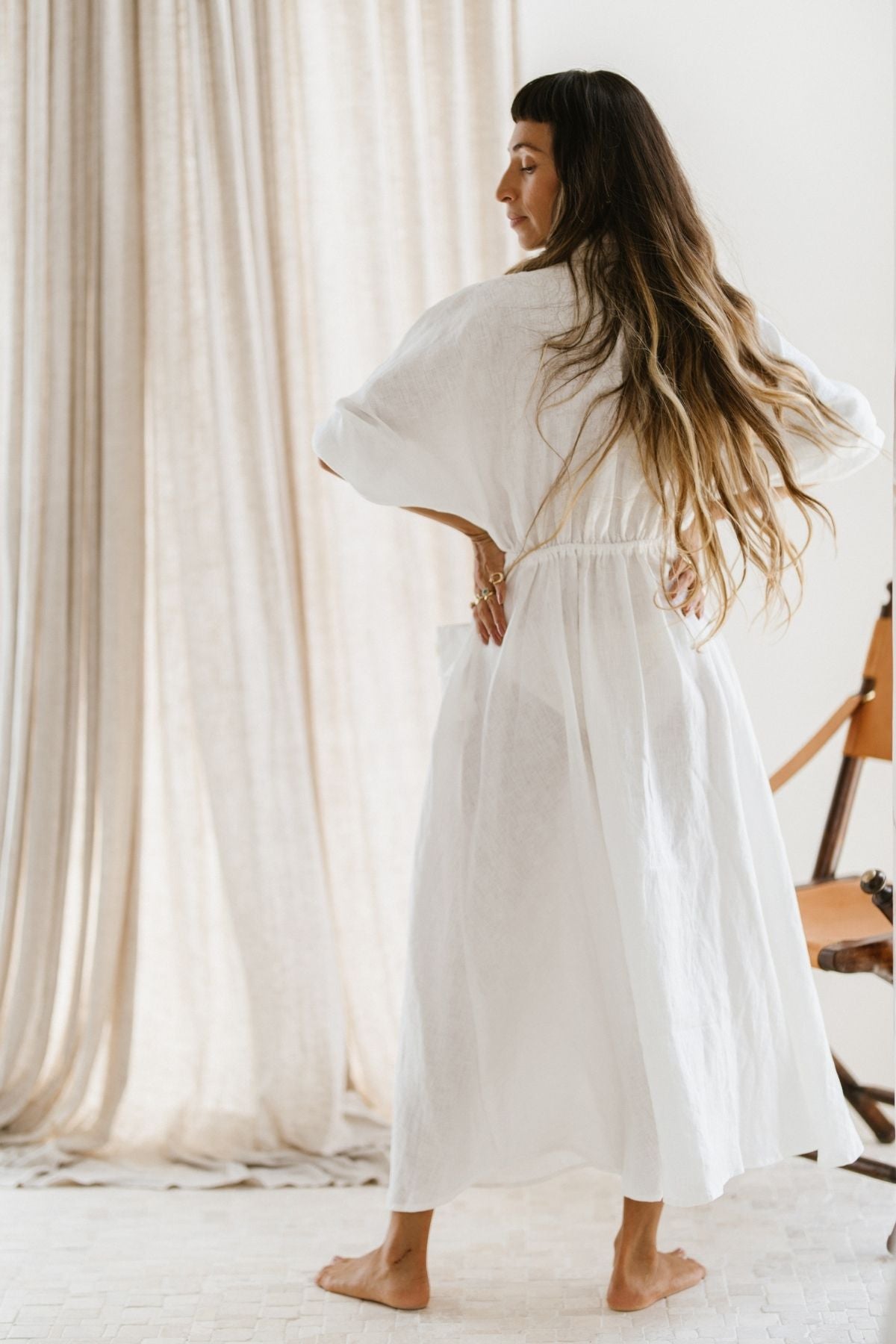 A woman with long hair stands barefoot on a light-colored floor, wearing the Kiki Long Dress (WI) by Myrah Penaloza, featuring a flowing white linen design with an adjustable waist. She is turned slightly to the side, hand on her hip, framed by a tall curtain and wooden furniture.
