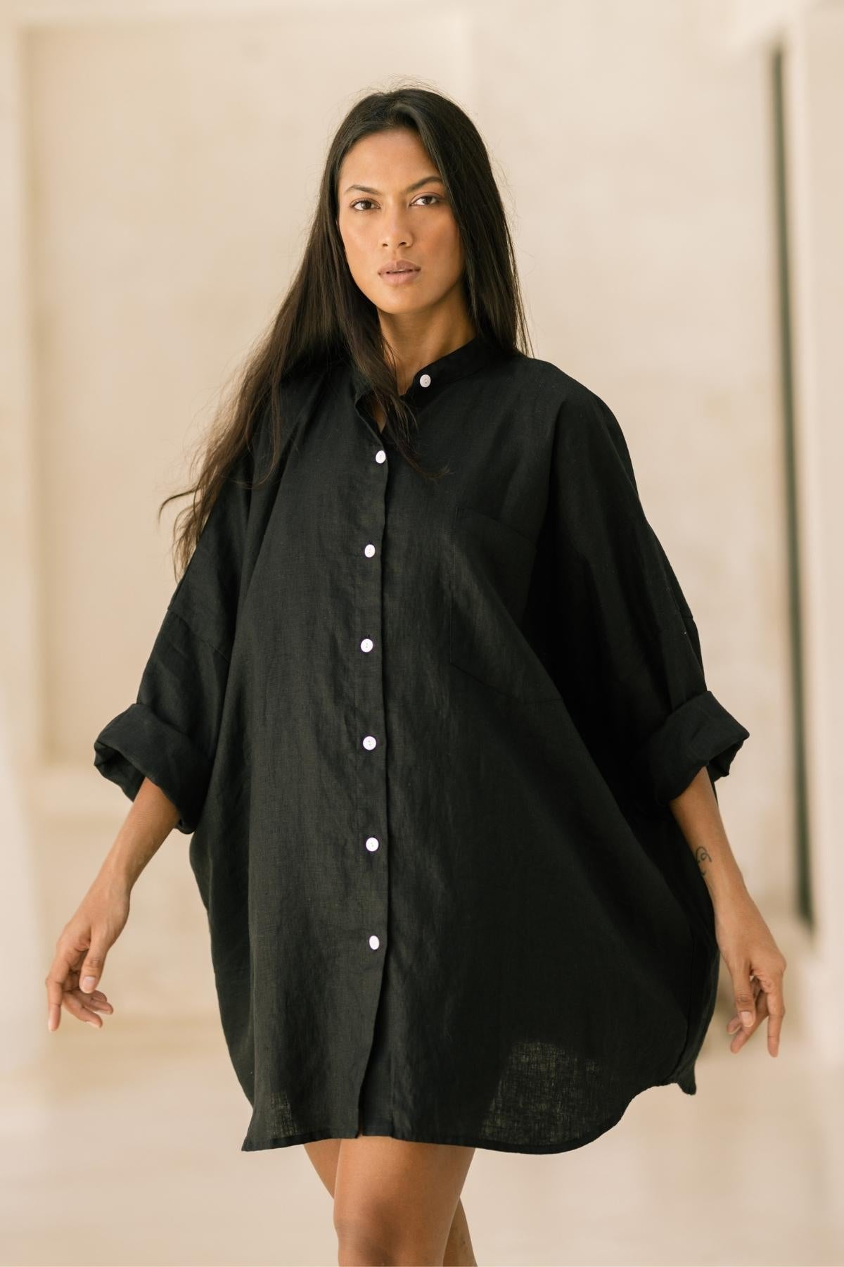 A woman with long, dark hair is wearing an oversized black Myrah Penaloza Camisa Del Novio Linen Top with the sleeves rolled up. She is standing in a light-colored, minimalist indoor setting, looking directly at the camera.