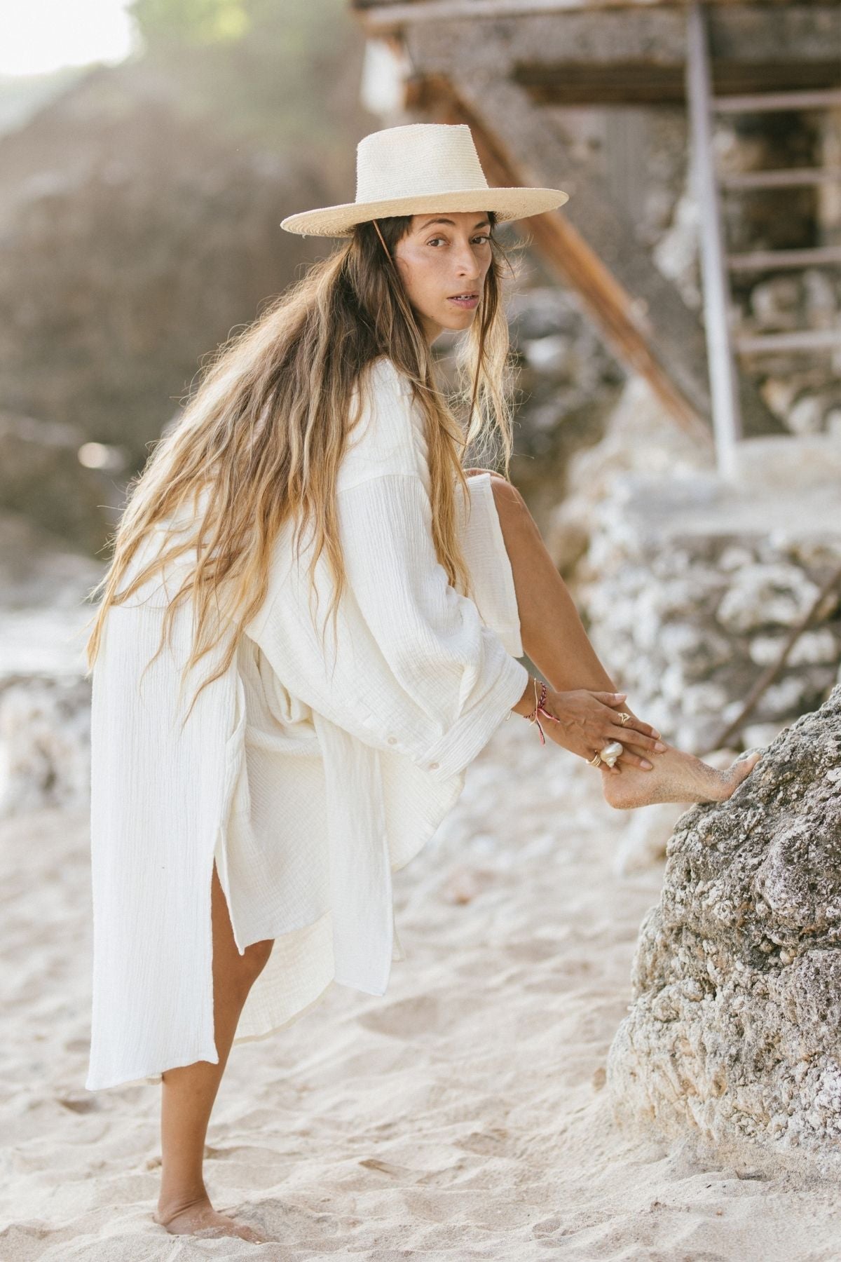 A woman in a Nidra Duster Organic Cotton / Hemp by Myrah Penaloza stands barefoot on a sandy beach, adjusting her foot on a rock, with a wooden beach hut in the background. The scene reflects relaxed, sustainable fashion in its purest form.