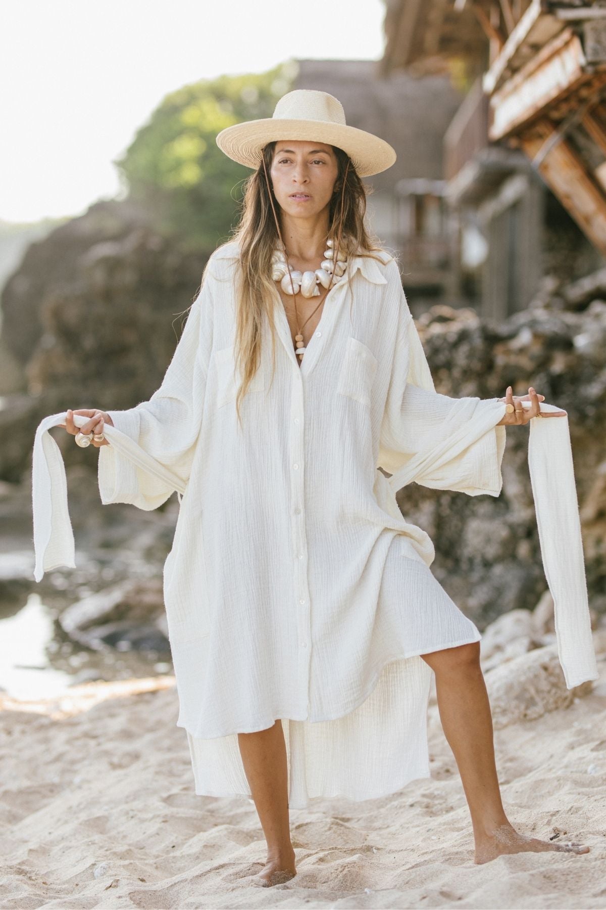 A person stands on a sandy beach wearing an oversized Nidra Duster from Myrah Penaloza, skillfully crafted from organic cotton and hemp. Paired with a straw hat, they hold onto the ties of the dress with a serene expression, as a rocky shoreline and lush greenery provide a picturesque backdrop.