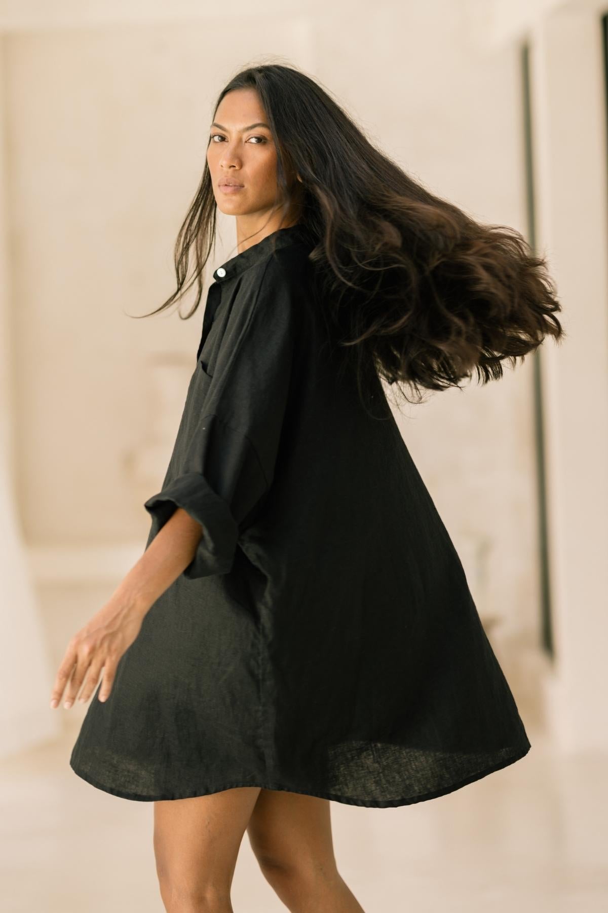 A woman with long, flowing hair is wearing the Camisa Del Novio Linen top paired with the Shanti Skirt from Myrah Penaloza. She is captured mid-turn, creating dynamic motion in her hair and outfit. The background is softly blurred, emphasizing her graceful movement in this elegant set.