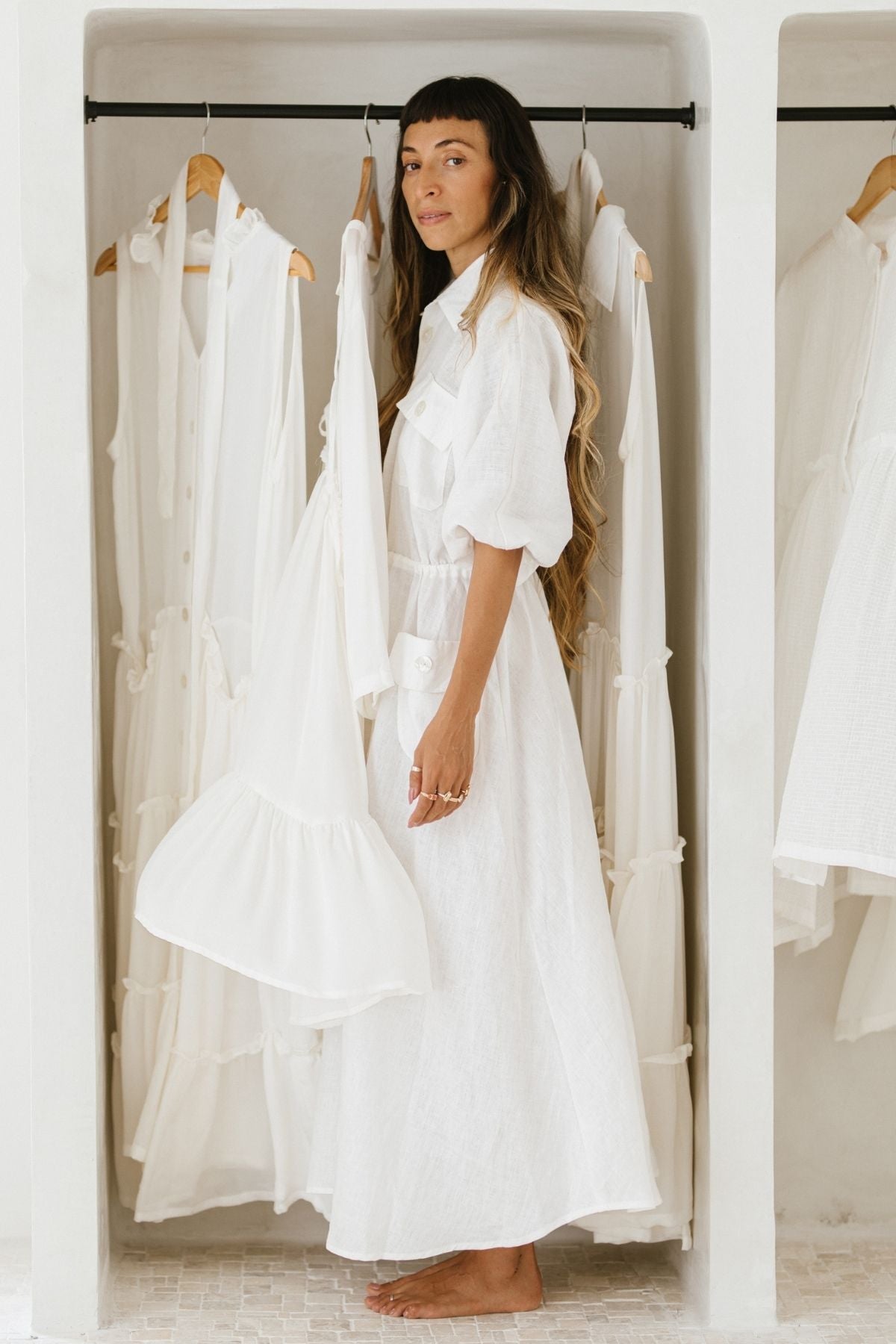 A woman with long hair stands barefoot in front of her wardrobe, elegantly draped in a Kiki Long Dress (WI) by Myrah Penaloza. Surrounding her are various oversized dresses on hangers, each designed with an adjustable waist. The setting is bright and minimalist.