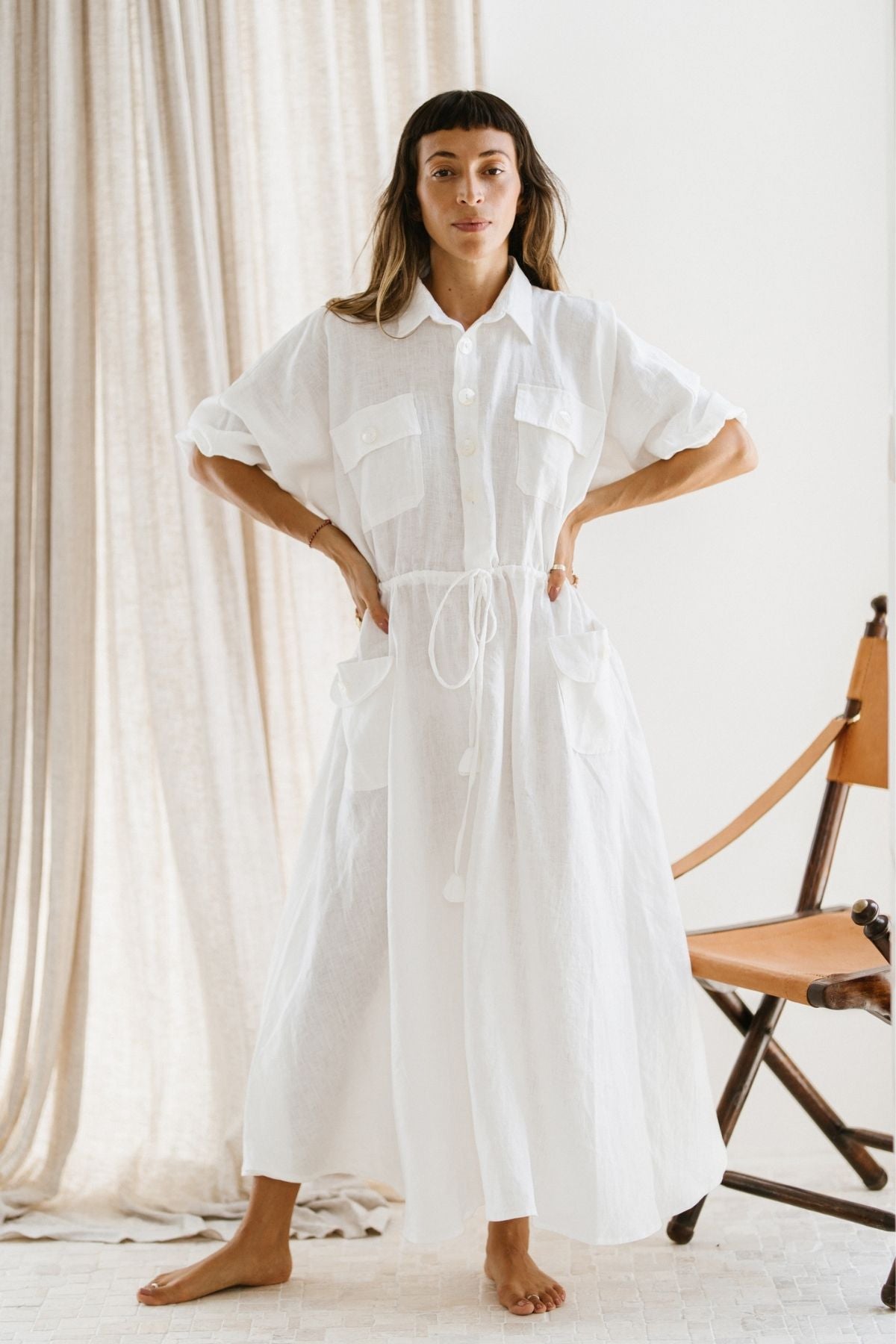 In a brightly lit room, an individual stands barefoot, dressed in the Kiki Long Dress (WI) by Myrah Penaloza. This oversized linen garment features a white button-up design with an adjustable waist. With hands on their hips, they are framed by a brown folding chair and beige curtains in the backdrop.