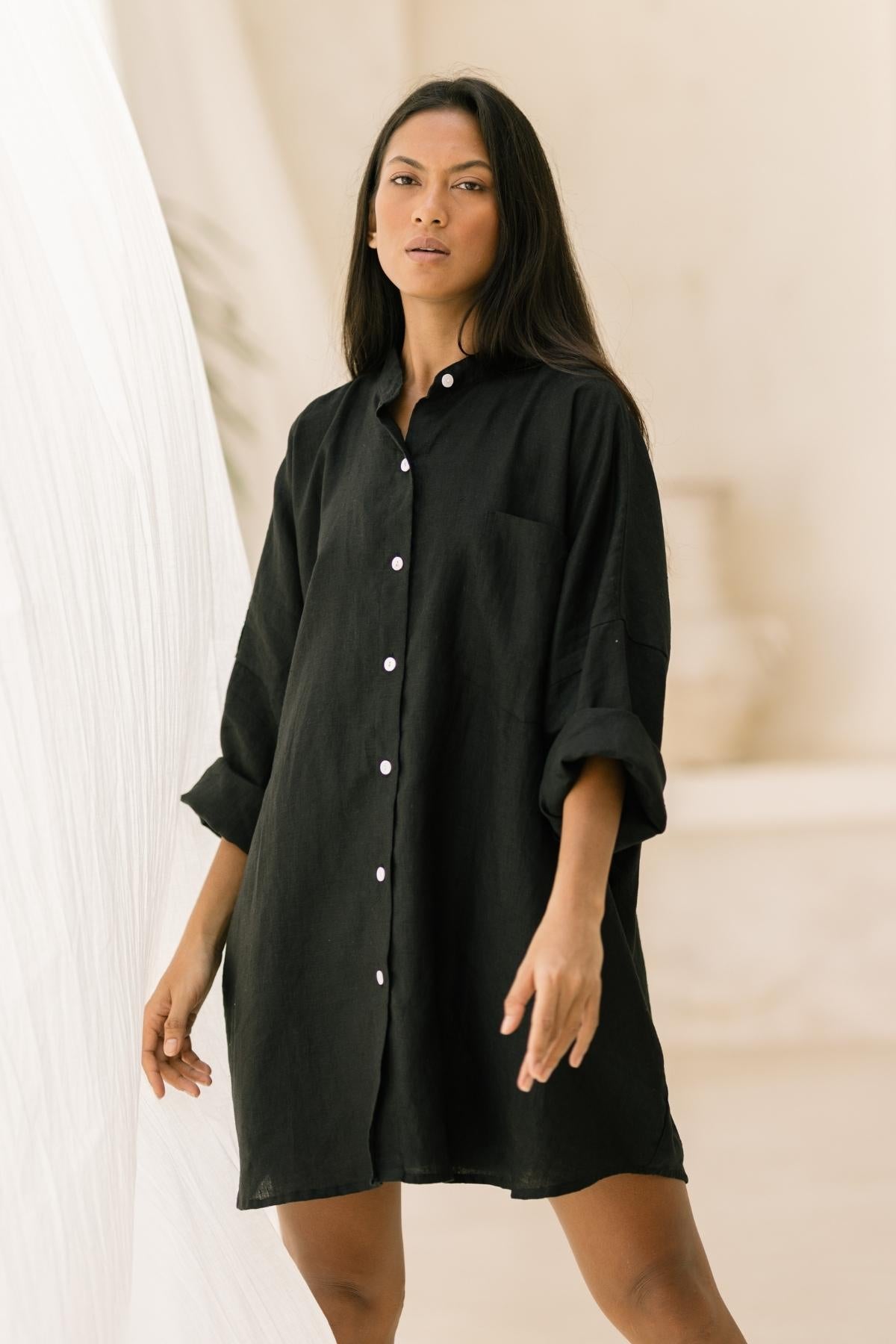 A person with long dark hair stands confidently in a relaxed black button-up shirt dress with rolled-up sleeves, evocative of the Camisa Del Novio Linen top from Myrah Penaloza, paired seamlessly with the Shanti Skirt. The background is softly blurred in neutral tones, featuring minimal decor elements.