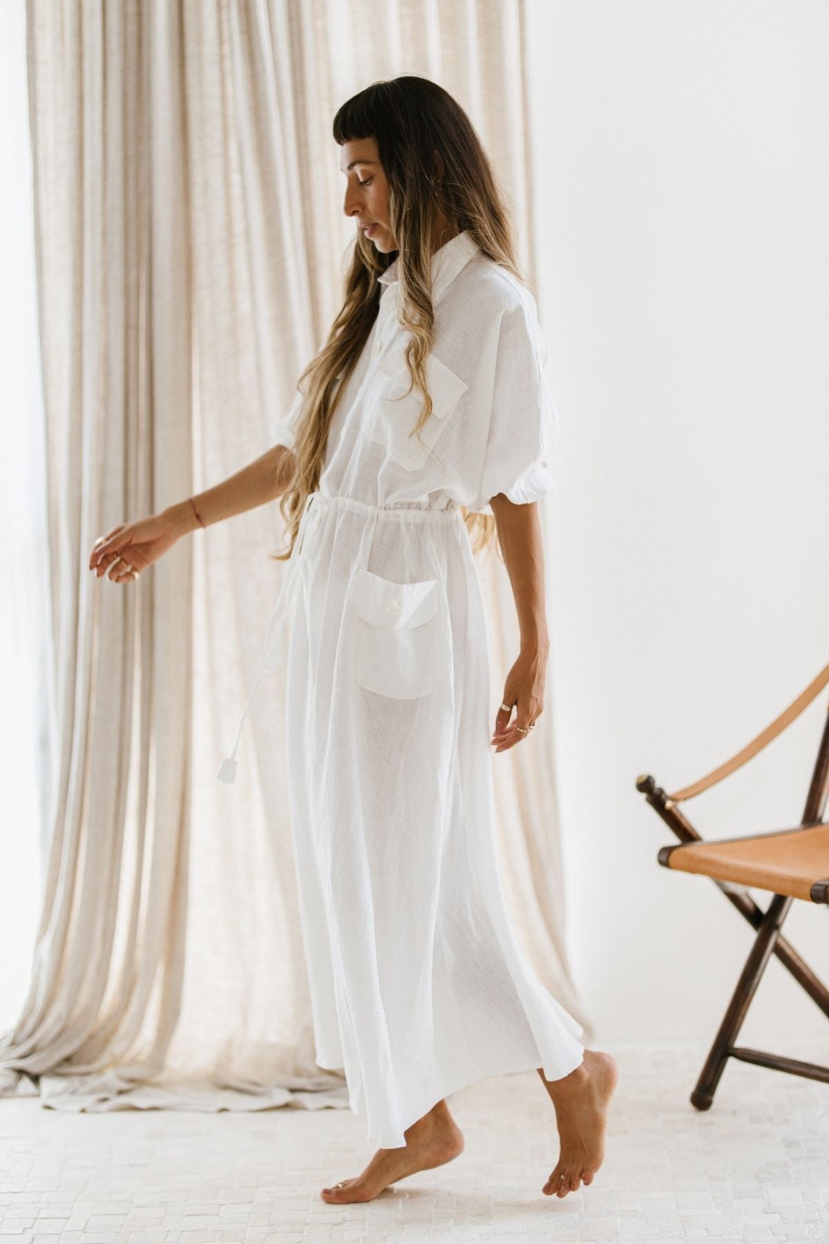 A woman with long hair is barefoot on a light floor, wearing the Kiki Long Dress (WI) by Myrah Penaloza, which features sheer white linen and an adjustable waist. She stands in front of beige curtains and a wooden chair with a light-colored seat, creating a minimal and bright setting.