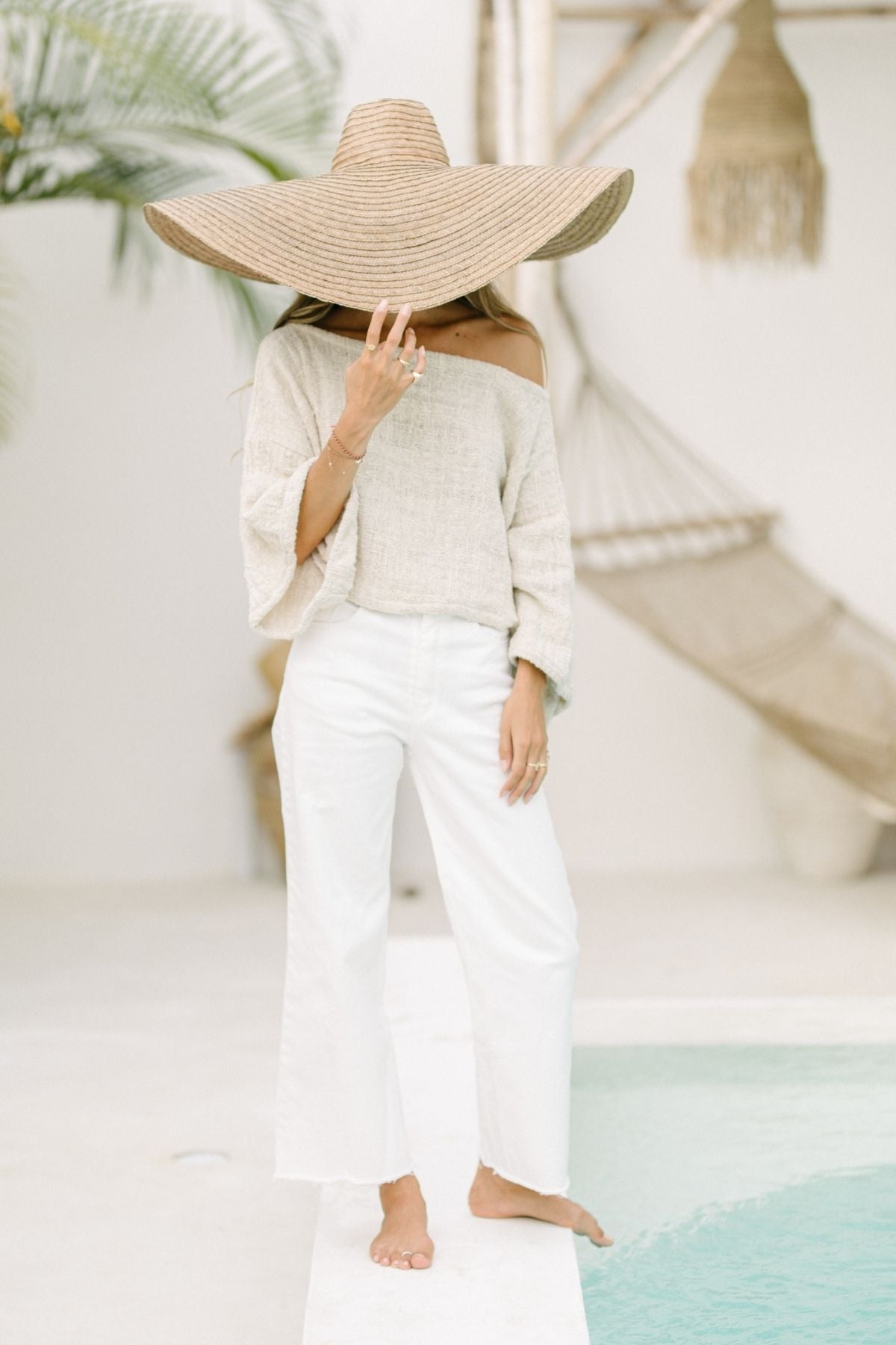 A woman stands near a pool, partially hiding her face with an oversized straw hat. She is wearing an off-the-shoulder, beige top with wide sleeves and the Miley (High Waisted) Jean by Myrah Penaloza. The background features tropical plants and a serene, light-colored decor.