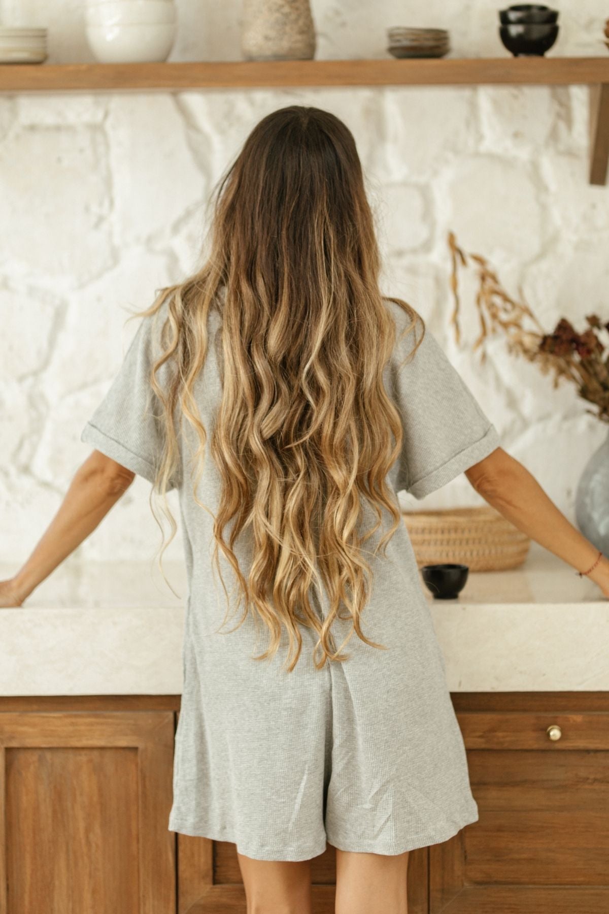 A woman with long, wavy blonde hair stands in front of a wooden countertop, facing away from the camera. She is wearing a gray Henley Playsuit by Myrah Penaloza. Behind her, a shelf displays various bowls and decorative items against a white textured wall, creating an ambiance perfect for cozy days.