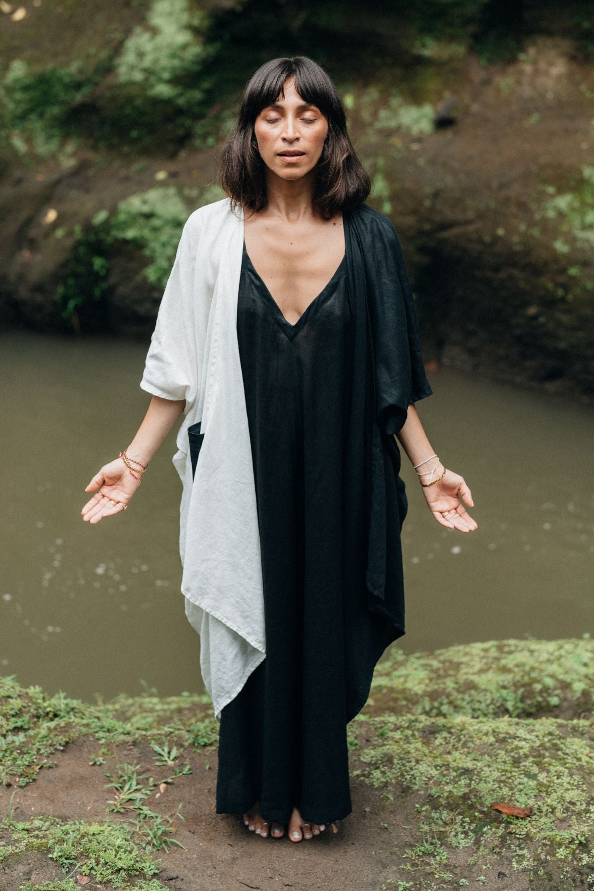 A woman stands in a meditative pose near a river, eyes closed, wearing the Dao Robe (Yin Yang) by Myrah Penaloza. She is barefoot, enveloped in lush greenery and the tranquility of nature.